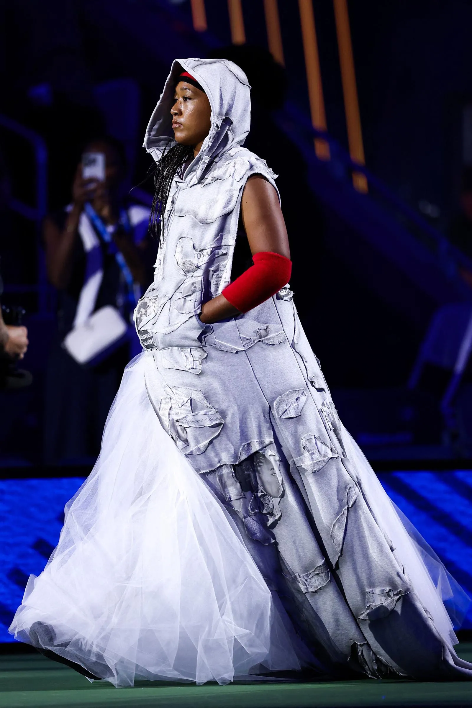 Person on a dim stage wearing a distressed gray hooded dress with a red armband on their left arm.