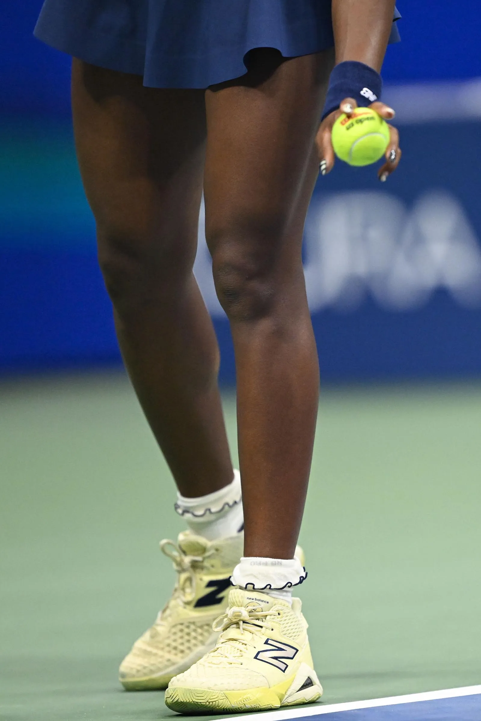 Close-up of a tennis player's legs and feet in cream sneakers on a court, gripping a bright yellow tennis ball in the right hand.