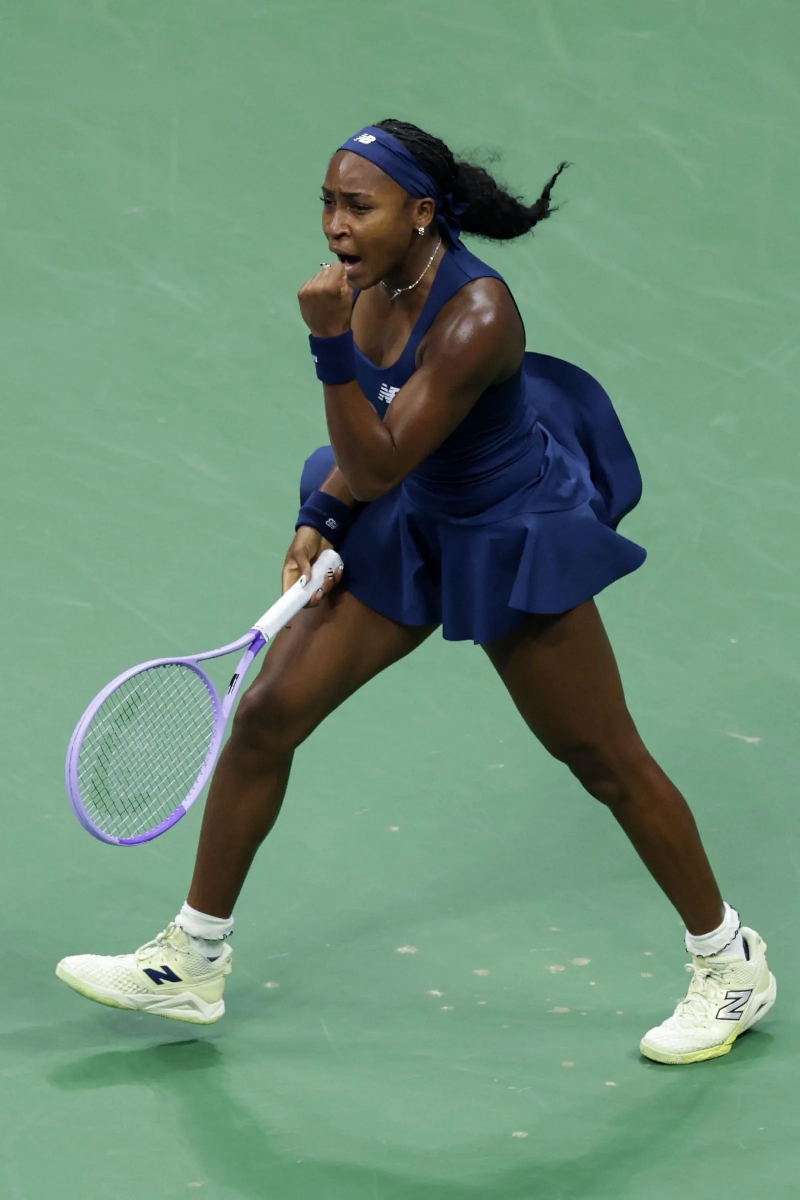 Female tennis player in a blue outfit mid-swing on a green court, holding a purple racket.