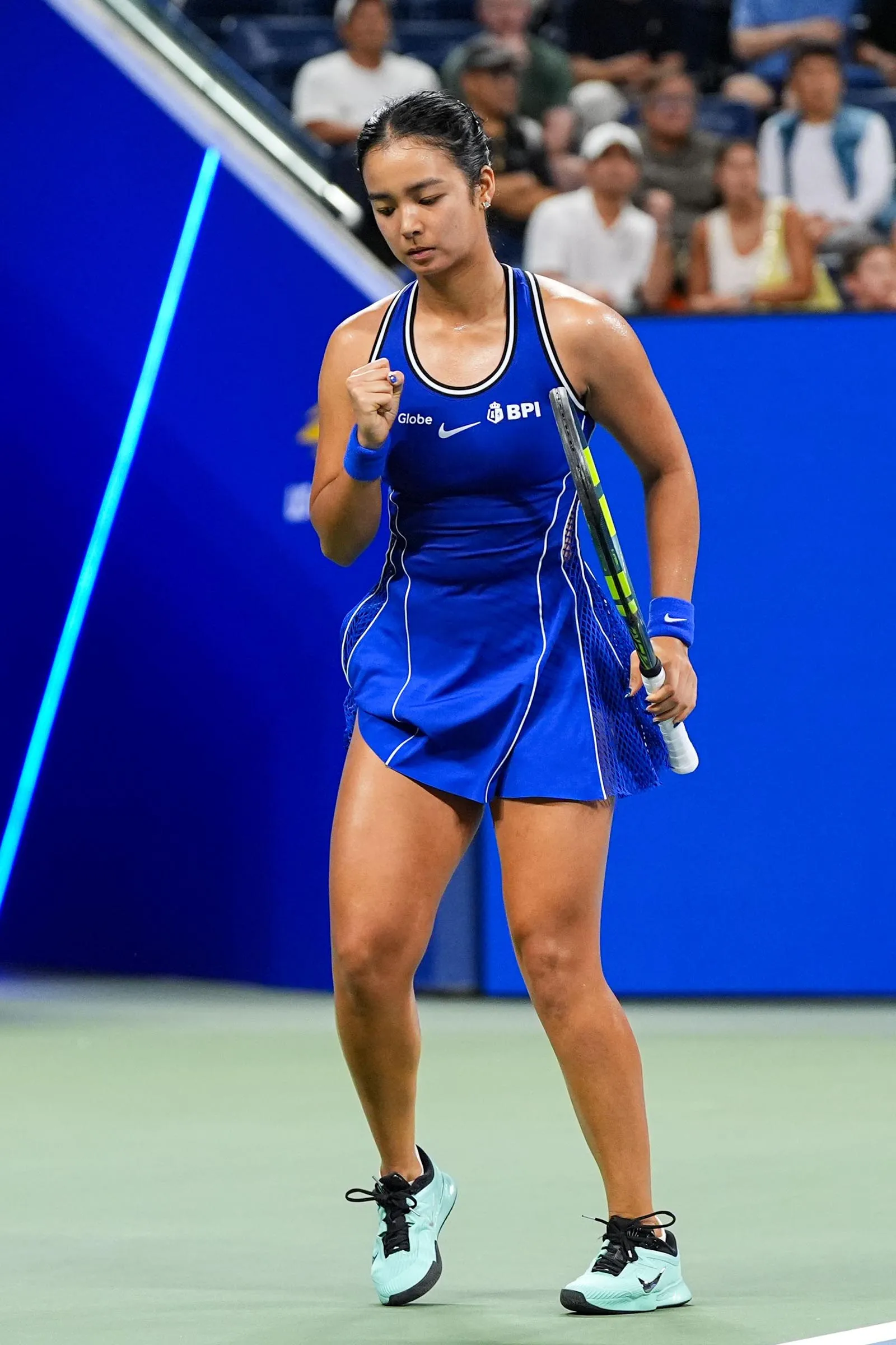 Female tennis player in a blue dress with white trim pumps her fist after a point on a tennis court, holding a racket in her other hand.