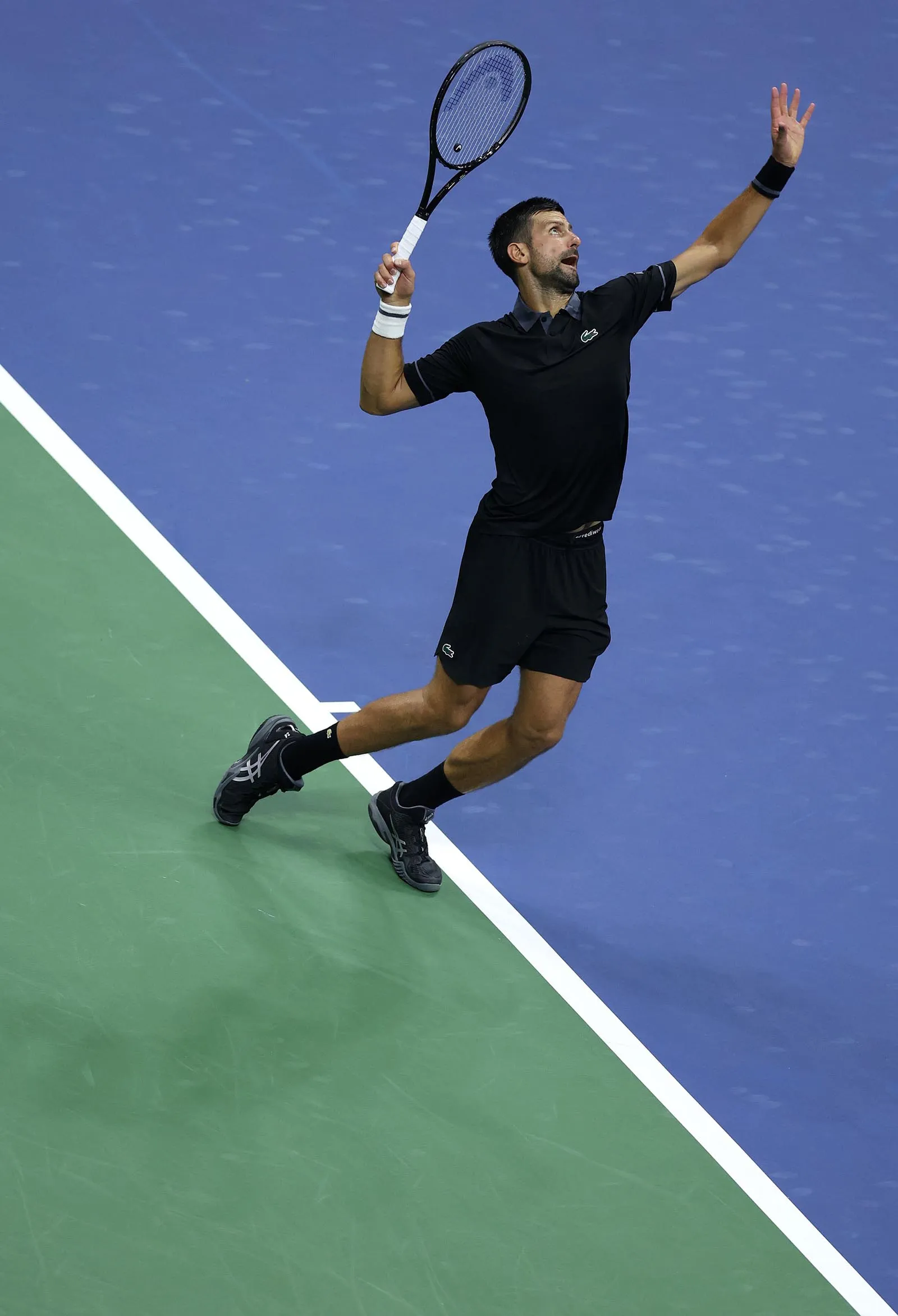 Male tennis player in black athletic wear leaps with racket raised on a blue court, preparing a shot as the green sideline shows nearby.