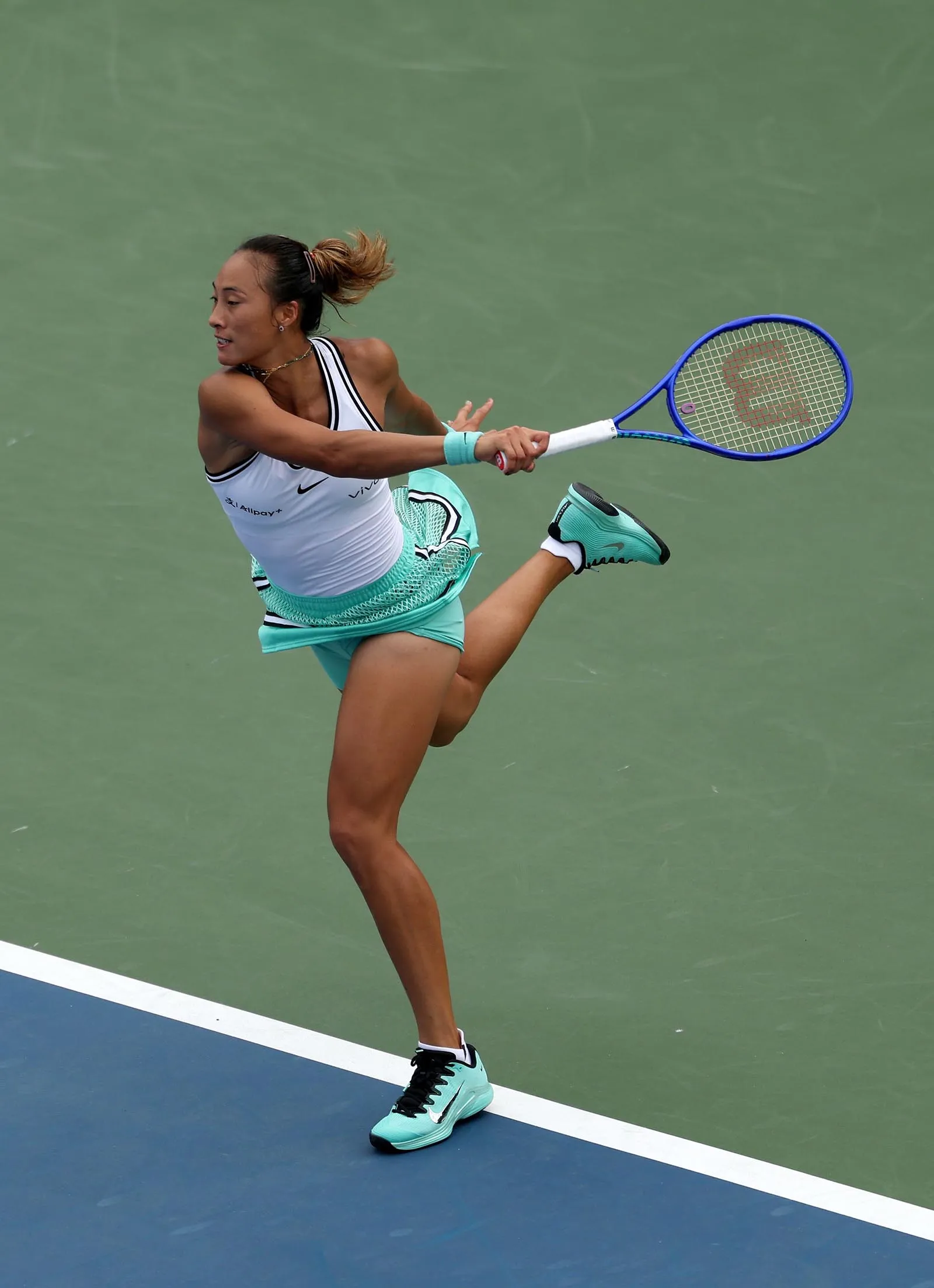 Female tennis player in mint-green outfit hitting a forehand while leaping on a court with a blue baseline and white line, blue racquet in motion.