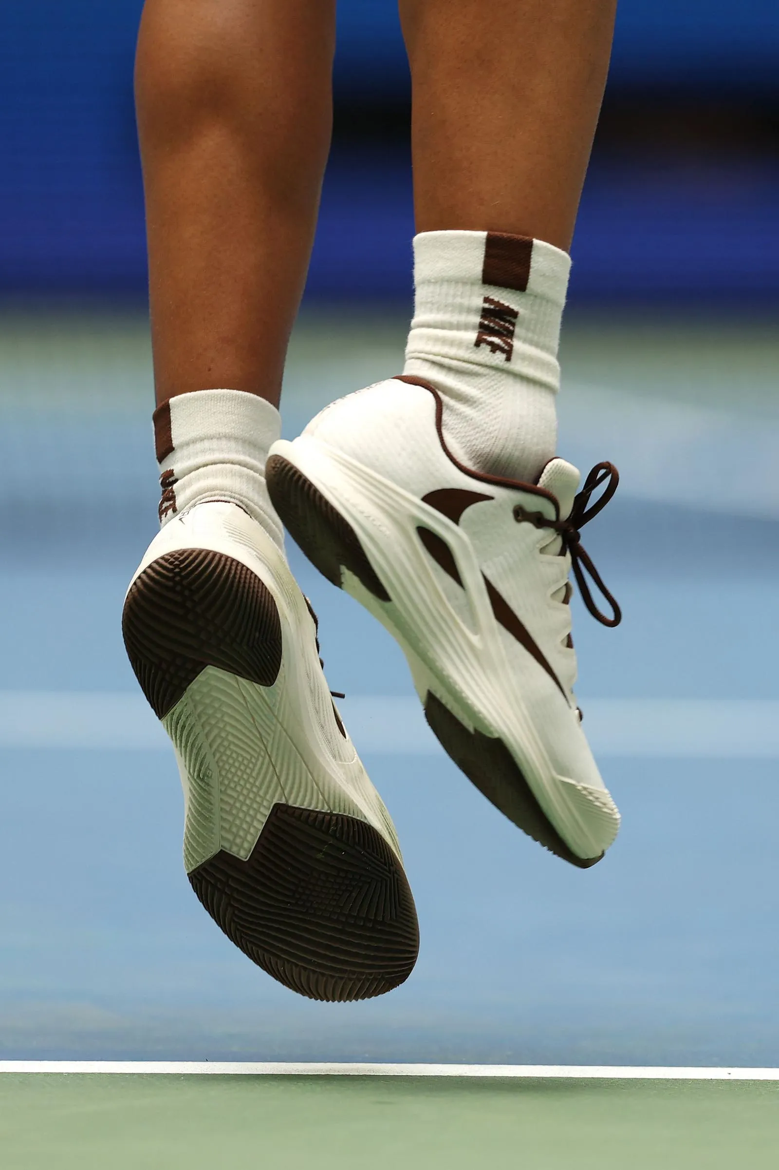Person's lower legs and white athletic sneakers with brown accents in mid-air over a tennis court, blue backdrop in background.