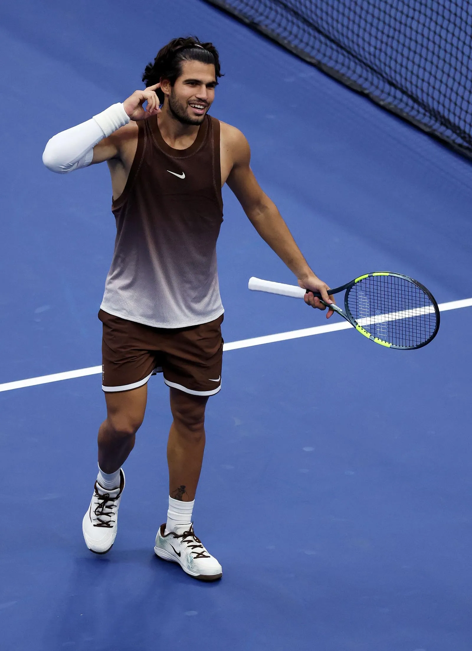 Tennis player on a blue court holding a racket in one hand, wearing a brown sleeveless top, brown shorts, and white socks and shoes, smiling and adjusting his ear with the other hand.