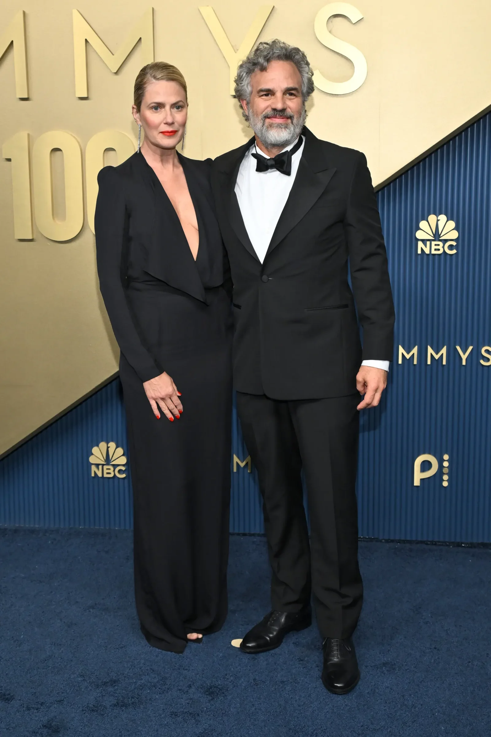 Man and woman in black formal attire posing together on a blue carpet at an NBC event, with a gold backdrop and NBC logos nearby.