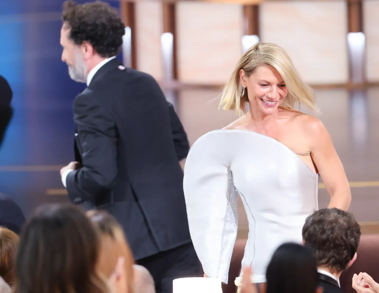 Smiling blonde woman in a white, off-shoulder gown walking through a crowded event as a man in a dark suit passes by behind her.