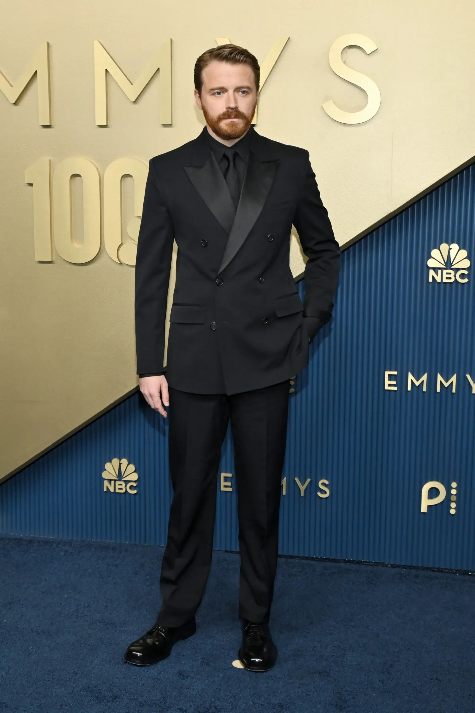 Man in a black double‑breasted suit with a black shirt and tie, standing with one hand in his pocket on a blue carpet at an Emmy event, NBC logos in the background.