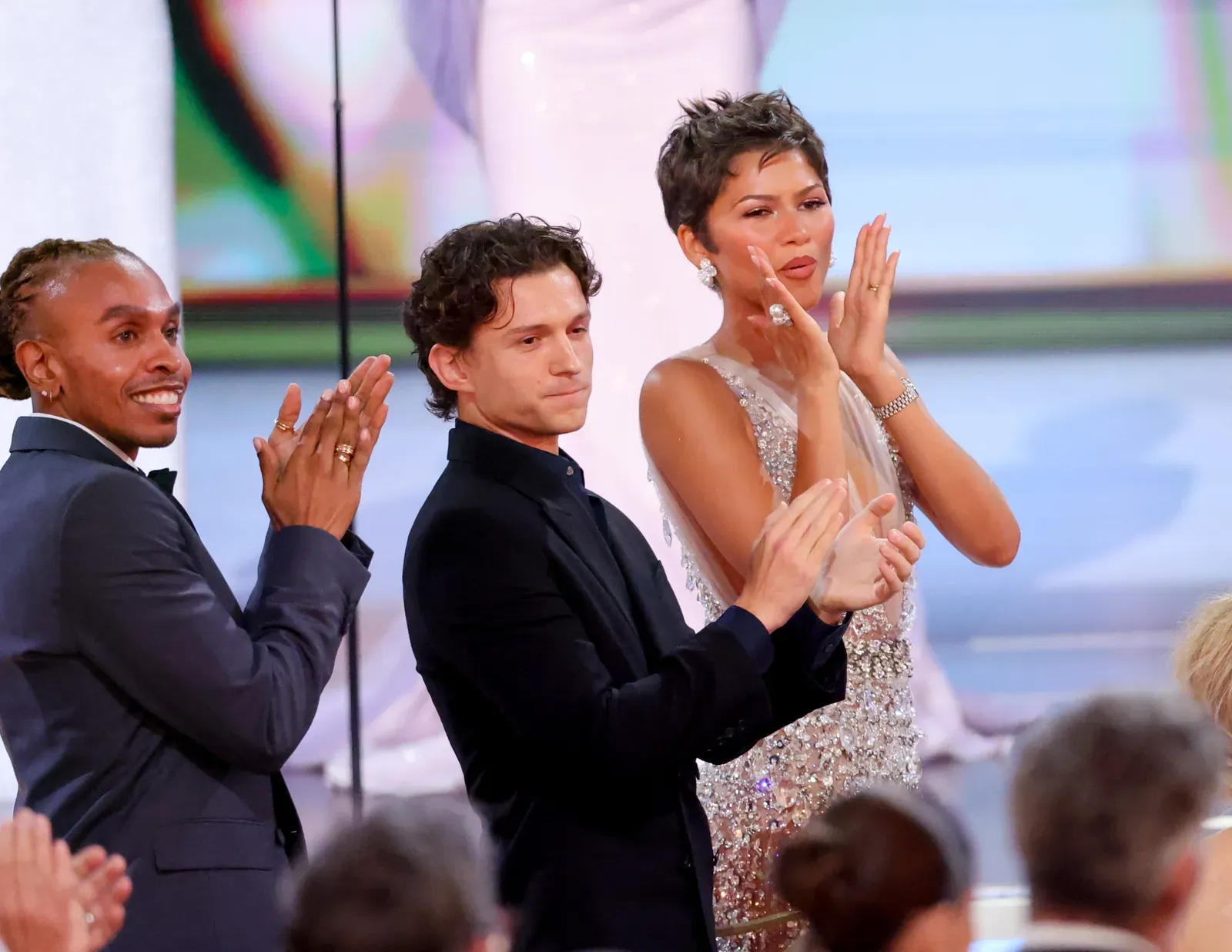Three formally dressed people clapping on a stage at an event with a crowd watching nearby.