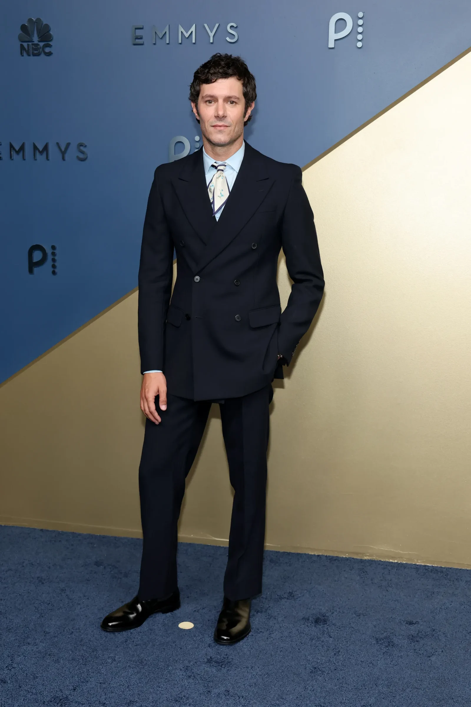 Full-length portrait of a man in a dark double-breasted suit and patterned tie standing on a blue-and-gold Emmy Awards backdrop at an event.