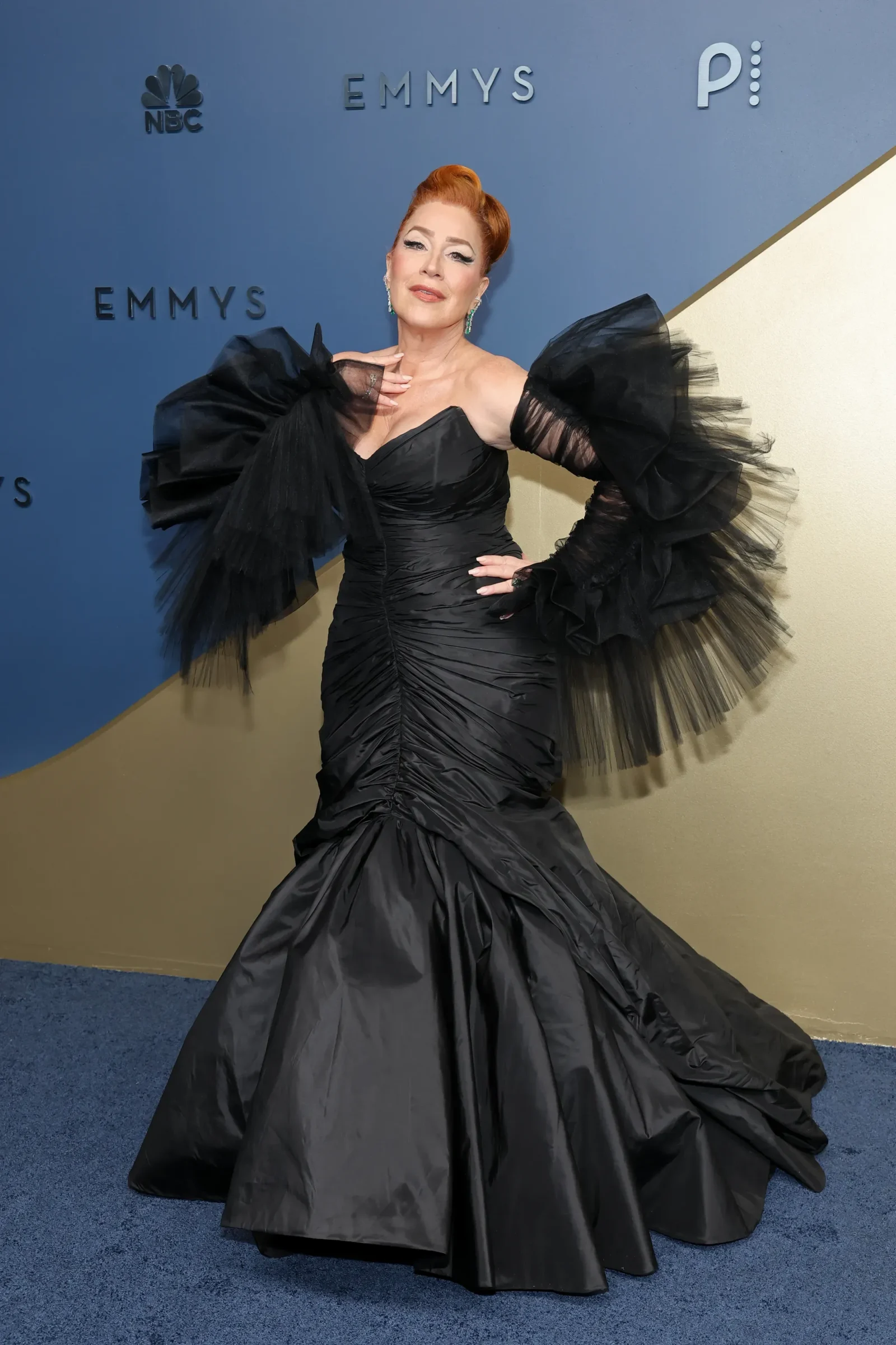 Woman in an elaborate black gown with oversized tulle sleeves posing on a blue Emmy Awards backdrop.