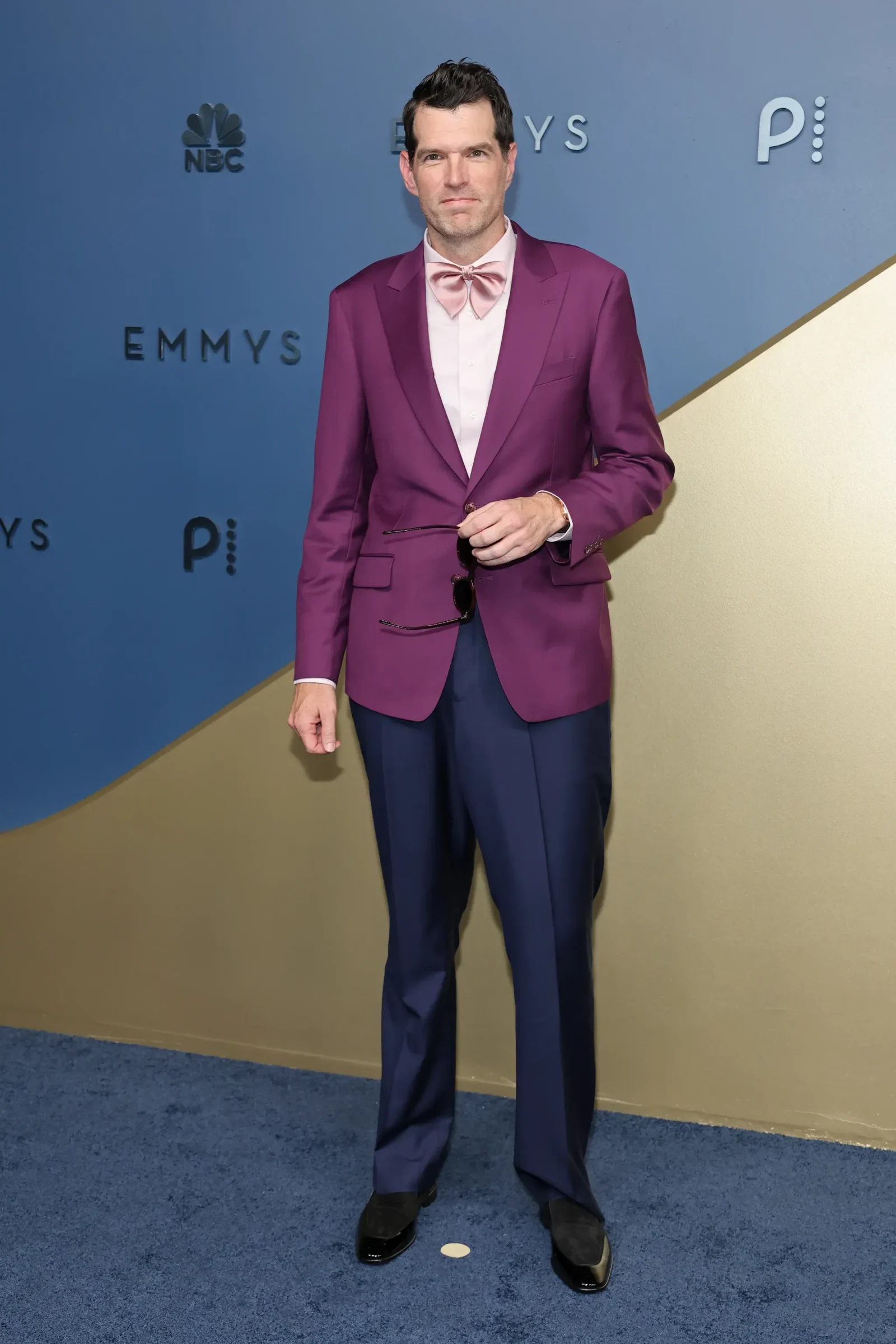 Full-length man in a purple suit with a pink bow tie posing on a blue Emmys backdrop, holding sunglasses in his hand.