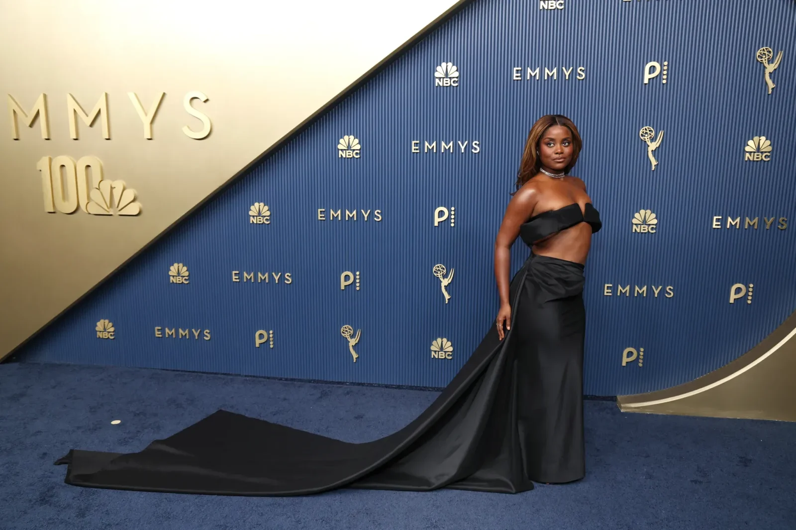 Woman in a black strapless gown posing on a blue Emmy Awards backdrop with long trailing train and jewelry