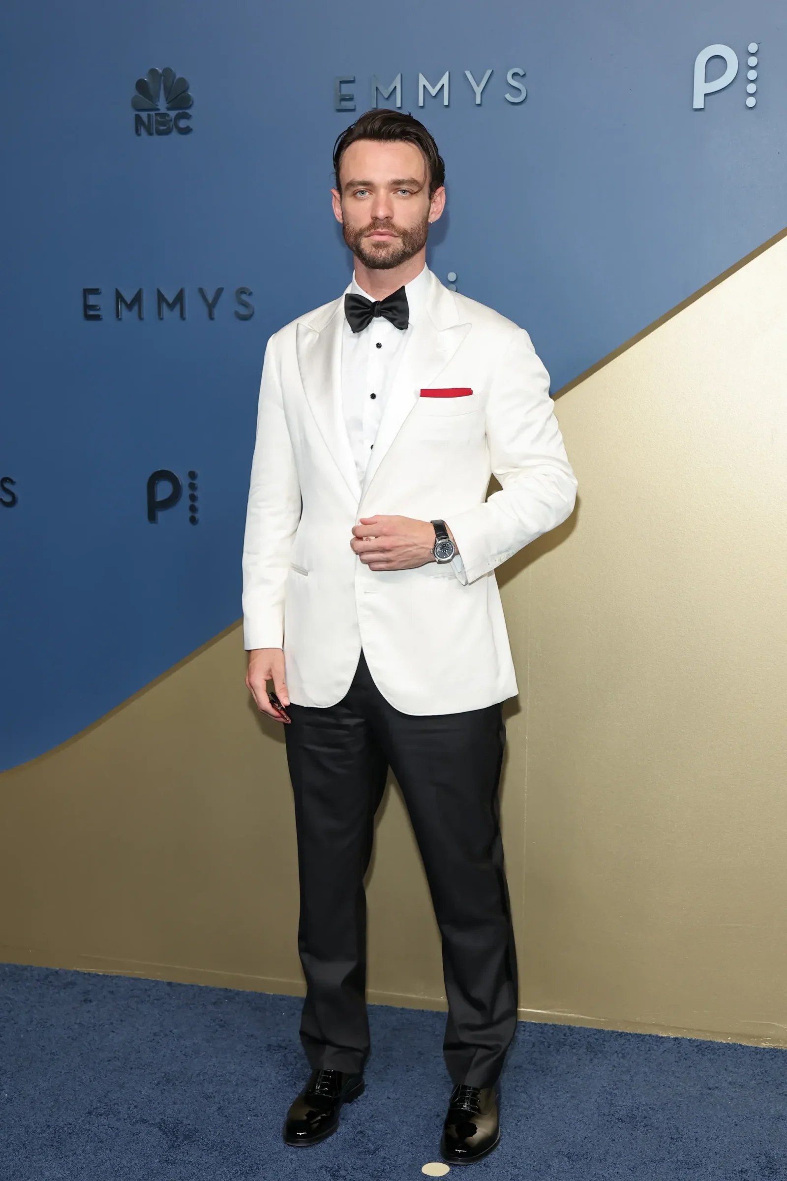 Male attendee in a white tuxedo with black bow tie, red pocket square, and black shoes posing on a blue Emmy backdrop.