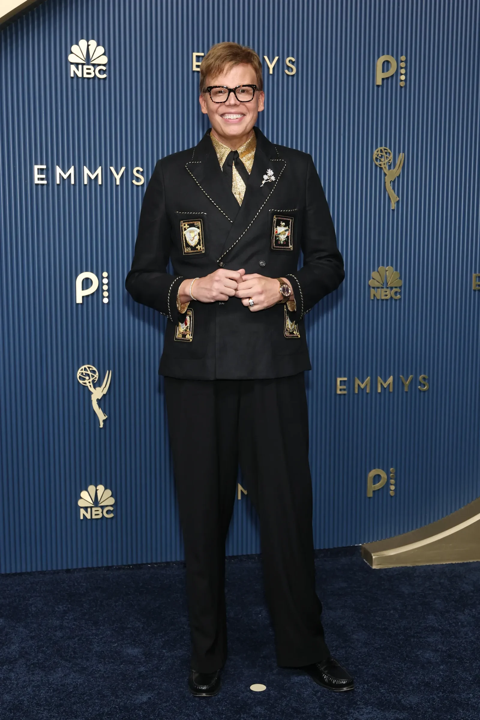 Person in a black suit with gold shirt and patches poses on Emmy backdrop with NBC logos, smiling at camera.