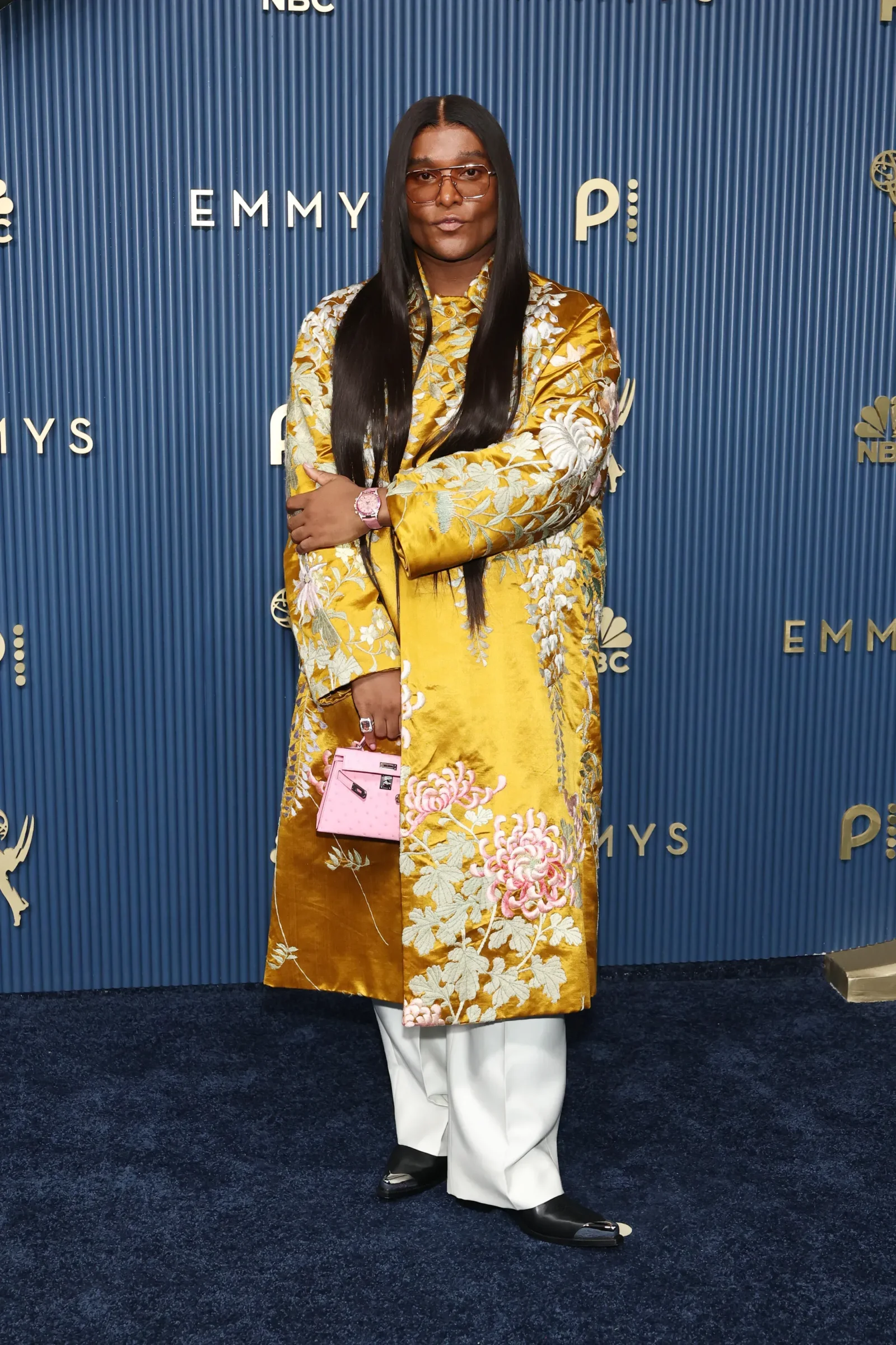 Person posing on a blue Emmy backdrop wearing a gold floral robe, long black hair, sunglasses, and a pink handbag.
