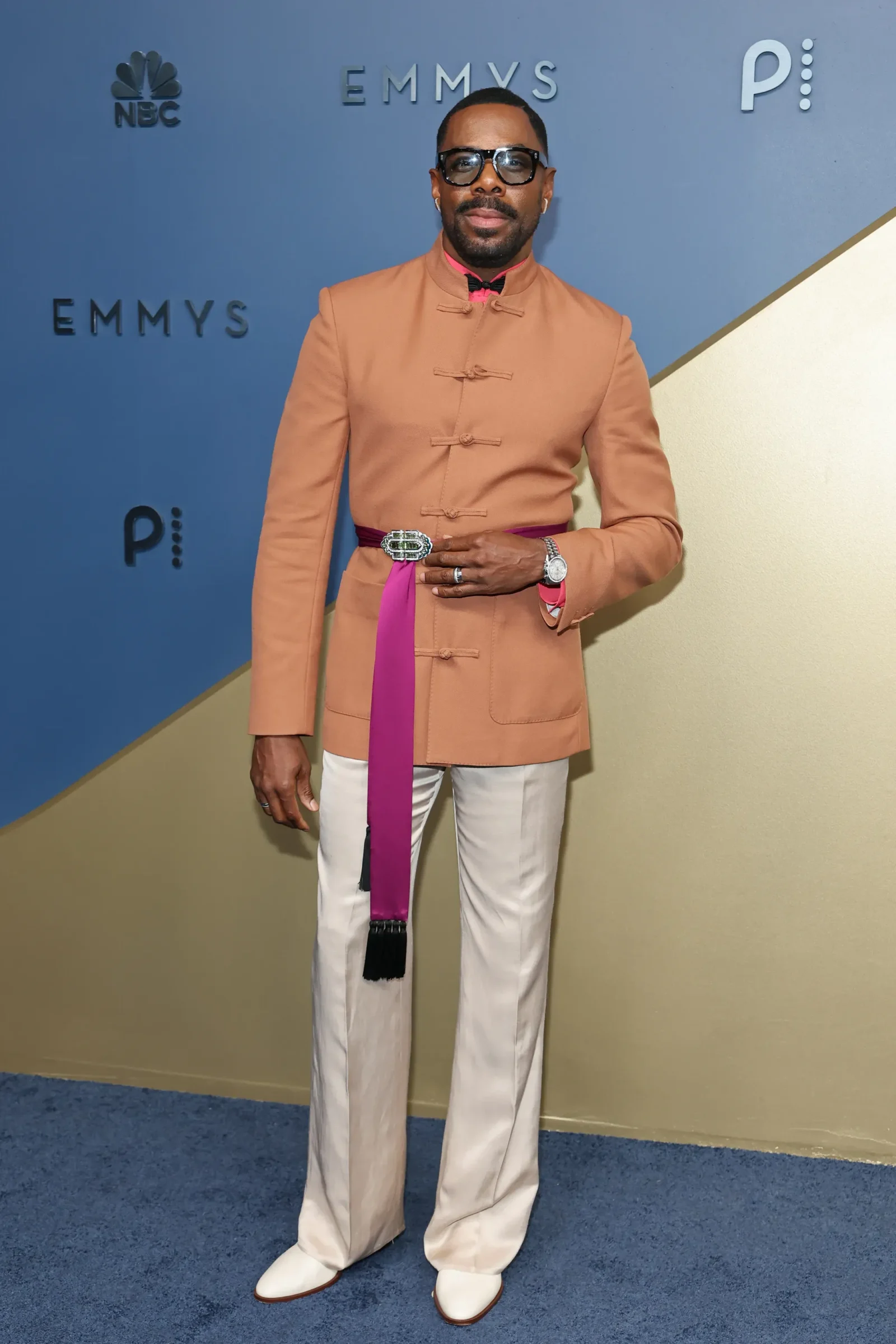 Man posing at an Emmy event wearing a tan Mandarin-collar jacket with a pink sash, white pants, and sunglasses against a blue backdrop with NBC logos.