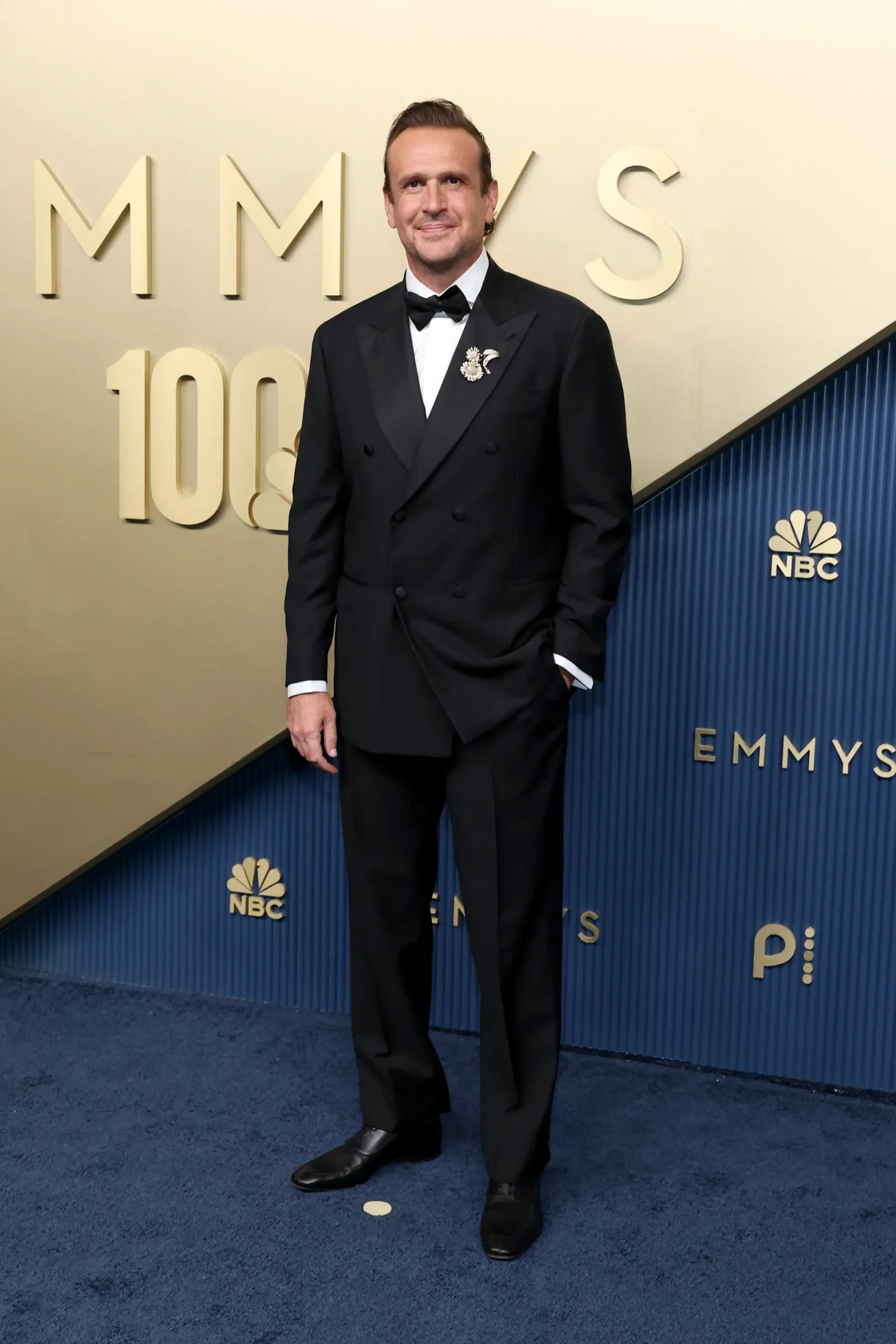 Man in a black tuxedo and bow tie posing on a beige Emmy Awards backdrop with NBC logos visible behind him, on blue carpeted floor.