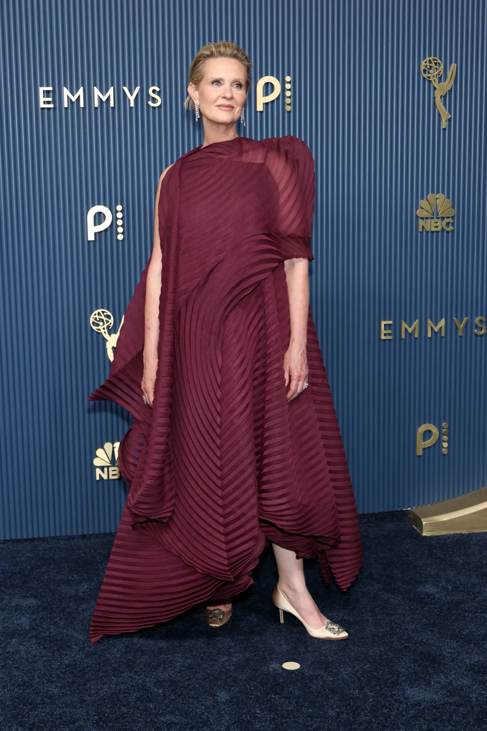 Woman posing on a blue Emmy backdrop in a burgundy pleated gown, accessorized with diamond earrings and nude heels.