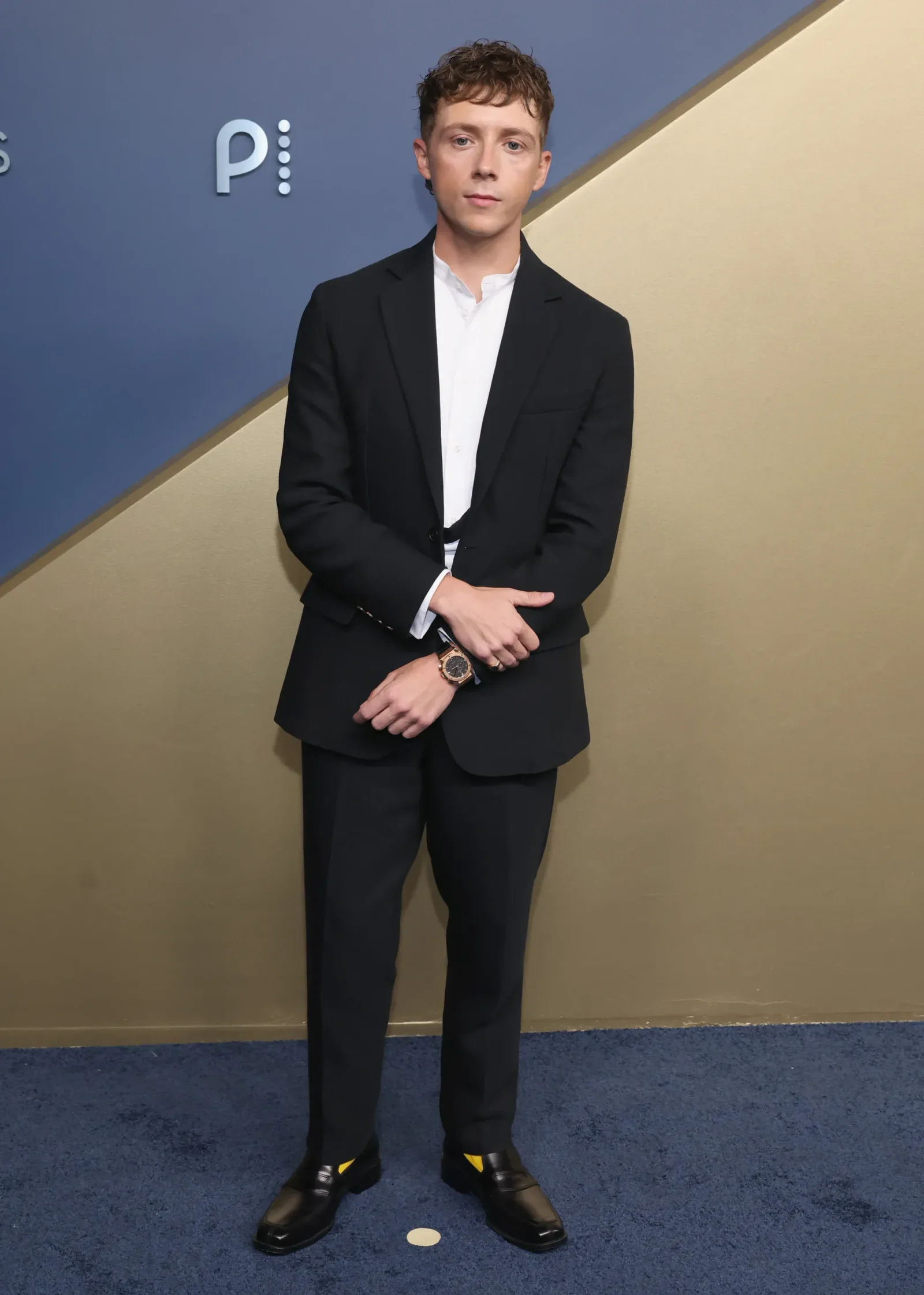Man in a black suit and white shirt standing against a blue and beige wall, hands folded in front.