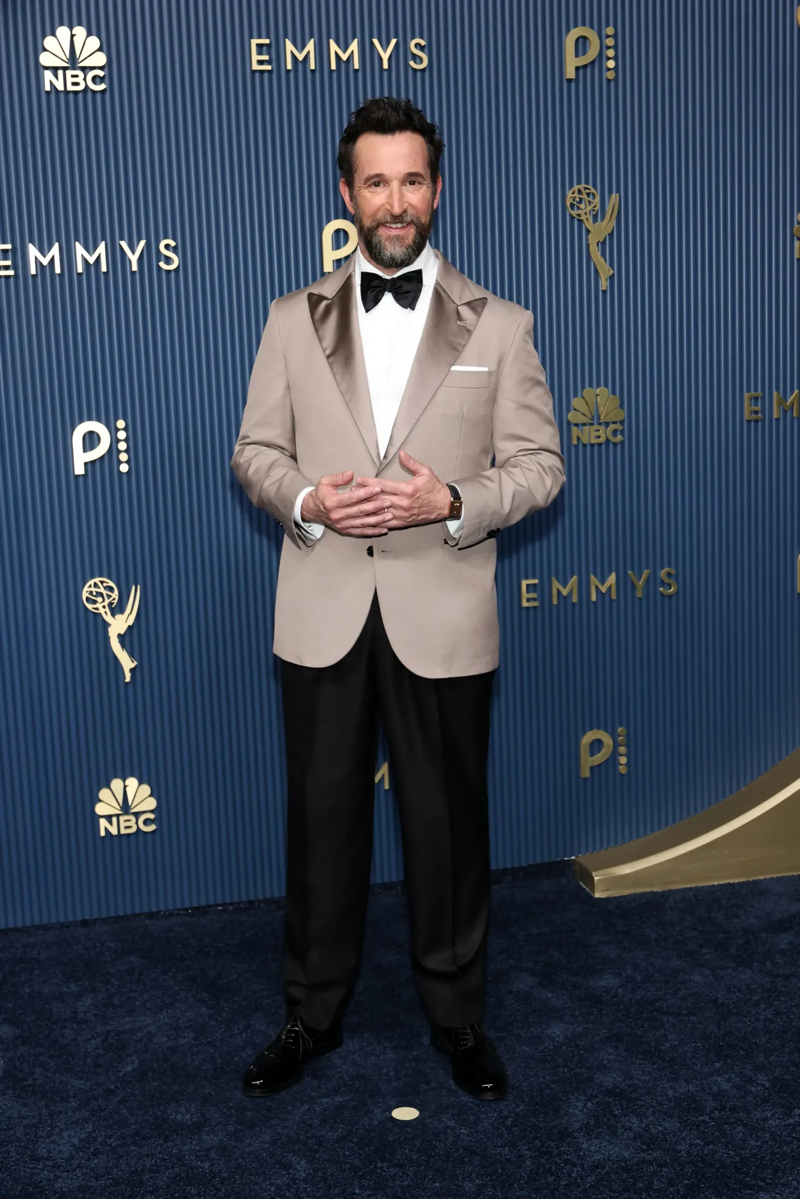 Man in a beige tuxedo with a black bow tie posing in front of a blue Emmy Awards backdrop with NBC logos.