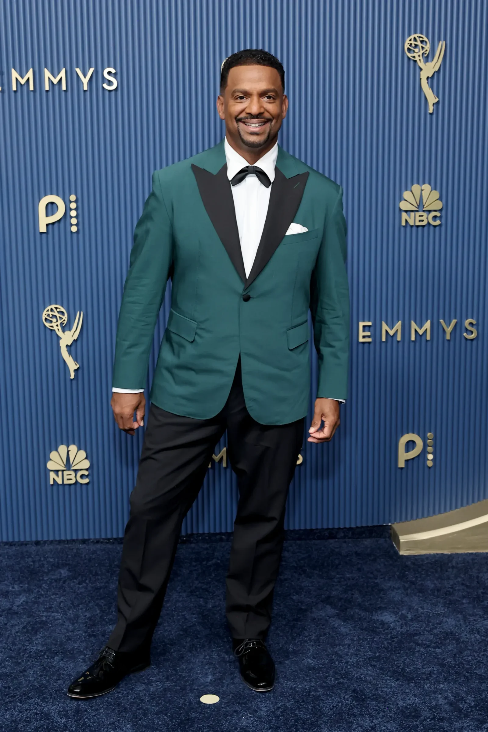 Man in a teal tuxedo with black lapels and bow tie posing on a blue Emmy Awards backdrop with NBC logos nearby