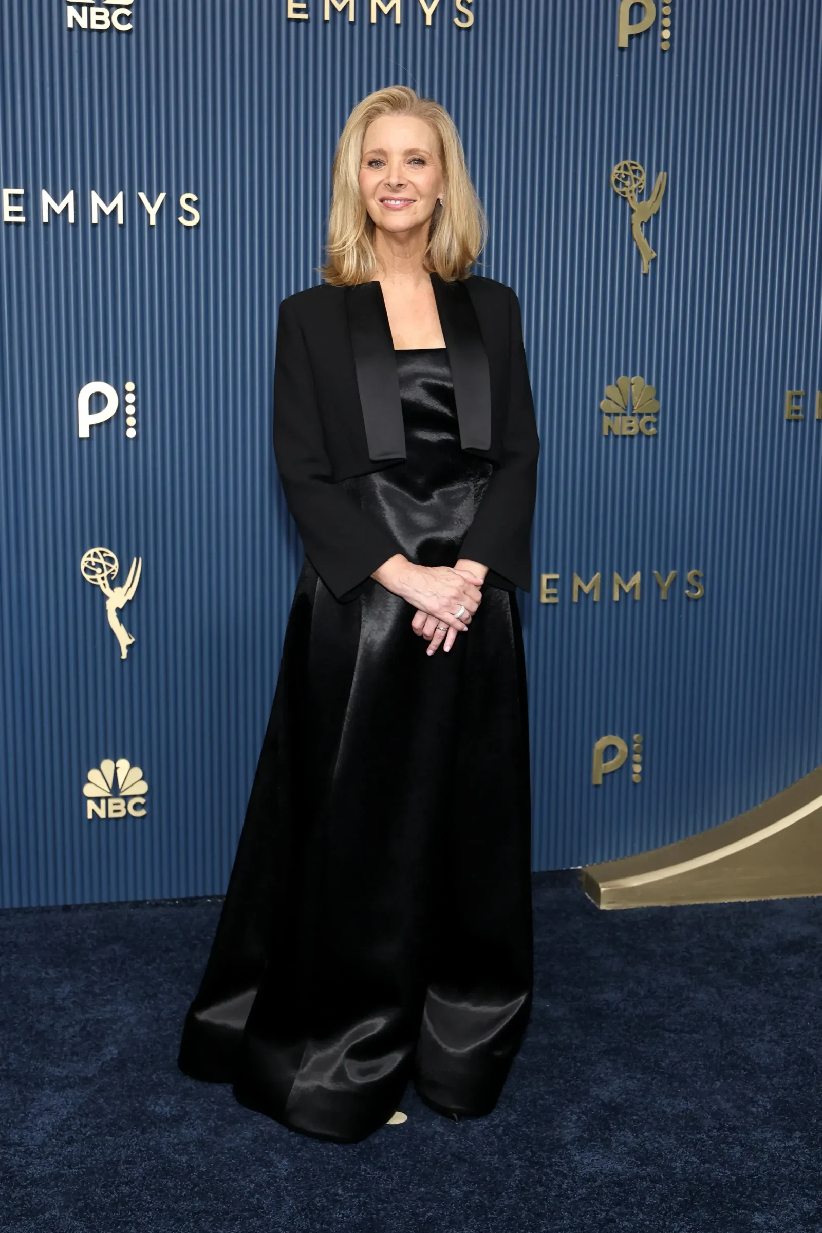Smiling blonde woman in a black satin gown poses on a blue Emmy Awards backdrop with logos behind her.