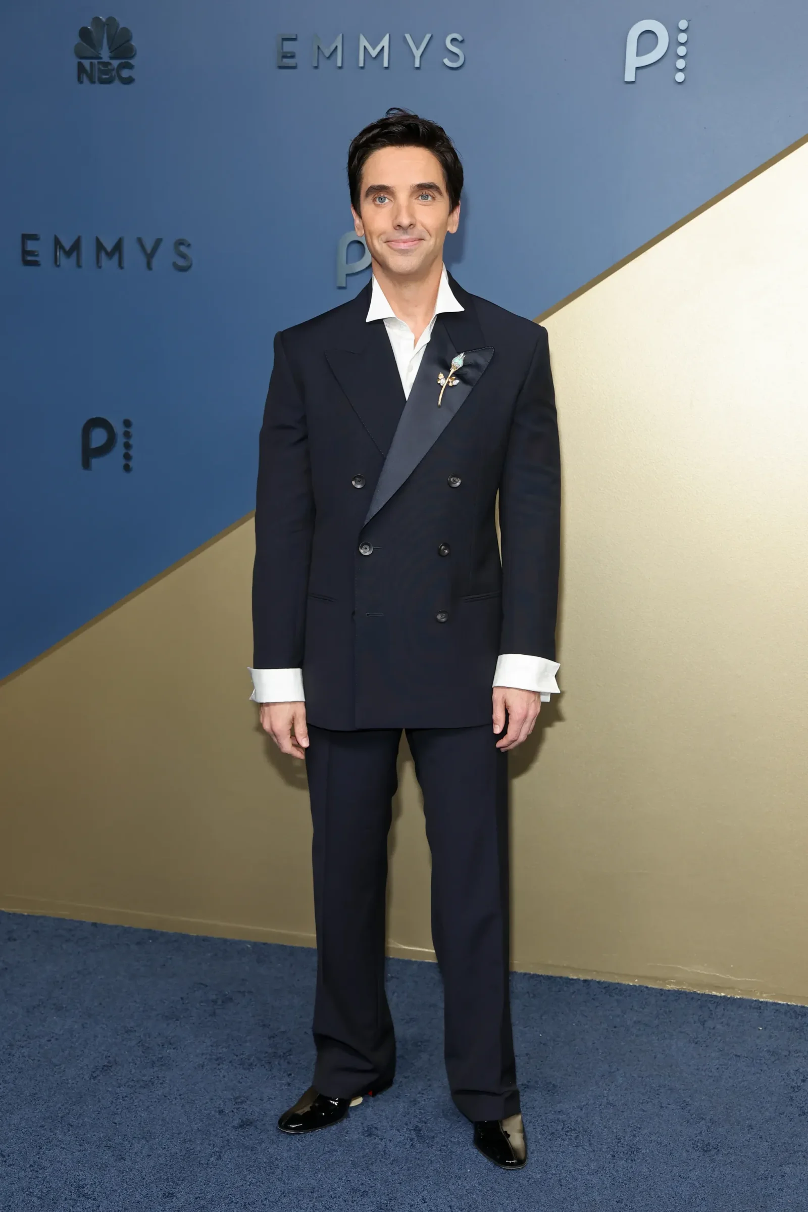 Man in a navy double-breasted suit with white shirt and boutonniere at an Emmy Awards backdrop.