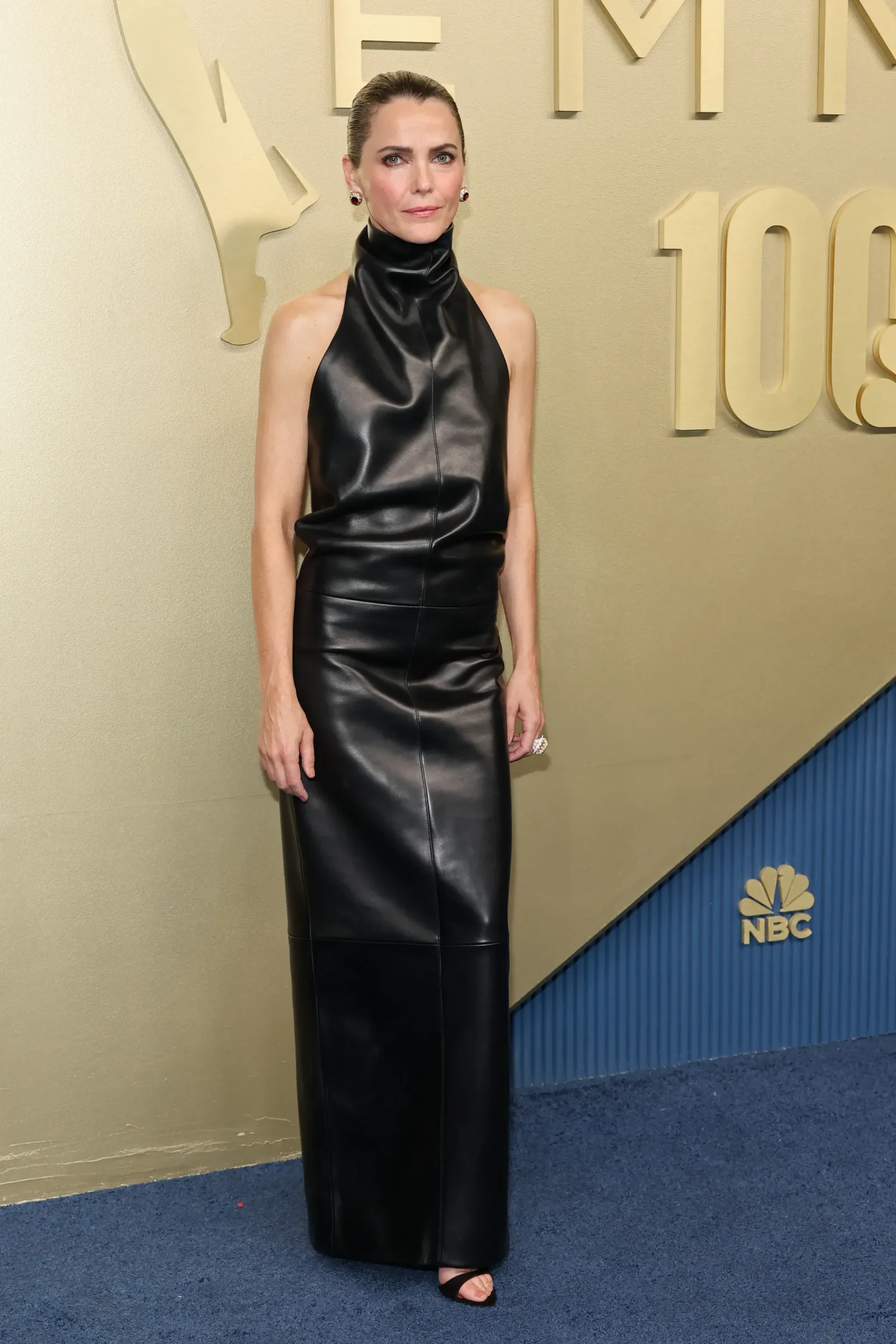 Full-length view of a woman in a black leather halter-neck dress posing on a gold NBC event backdrop with logo nearby.