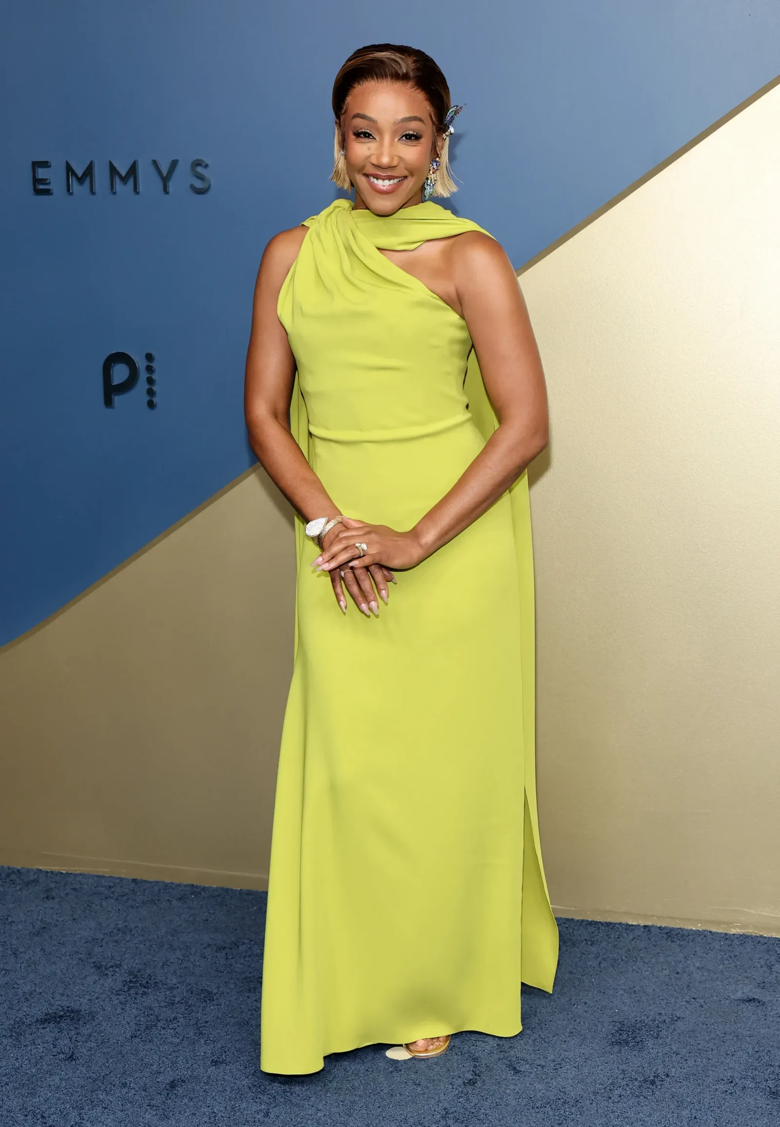 Smiling woman in a lime-green one-shoulder gown posing on a blue and beige backdrop at the Emmy Awards.