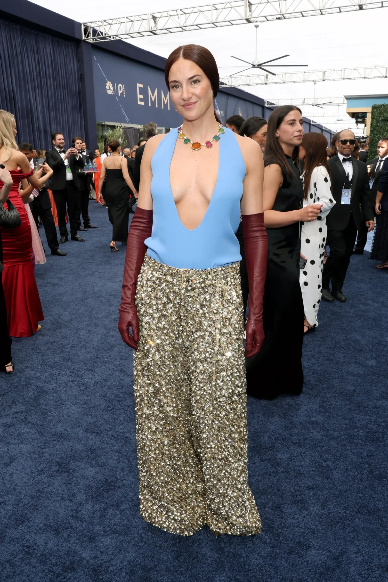 Woman on a blue carpet wearing a plunging light-blue top, a gold sequin skirt, burgundy gloves, and colorful necklace at a formal event.