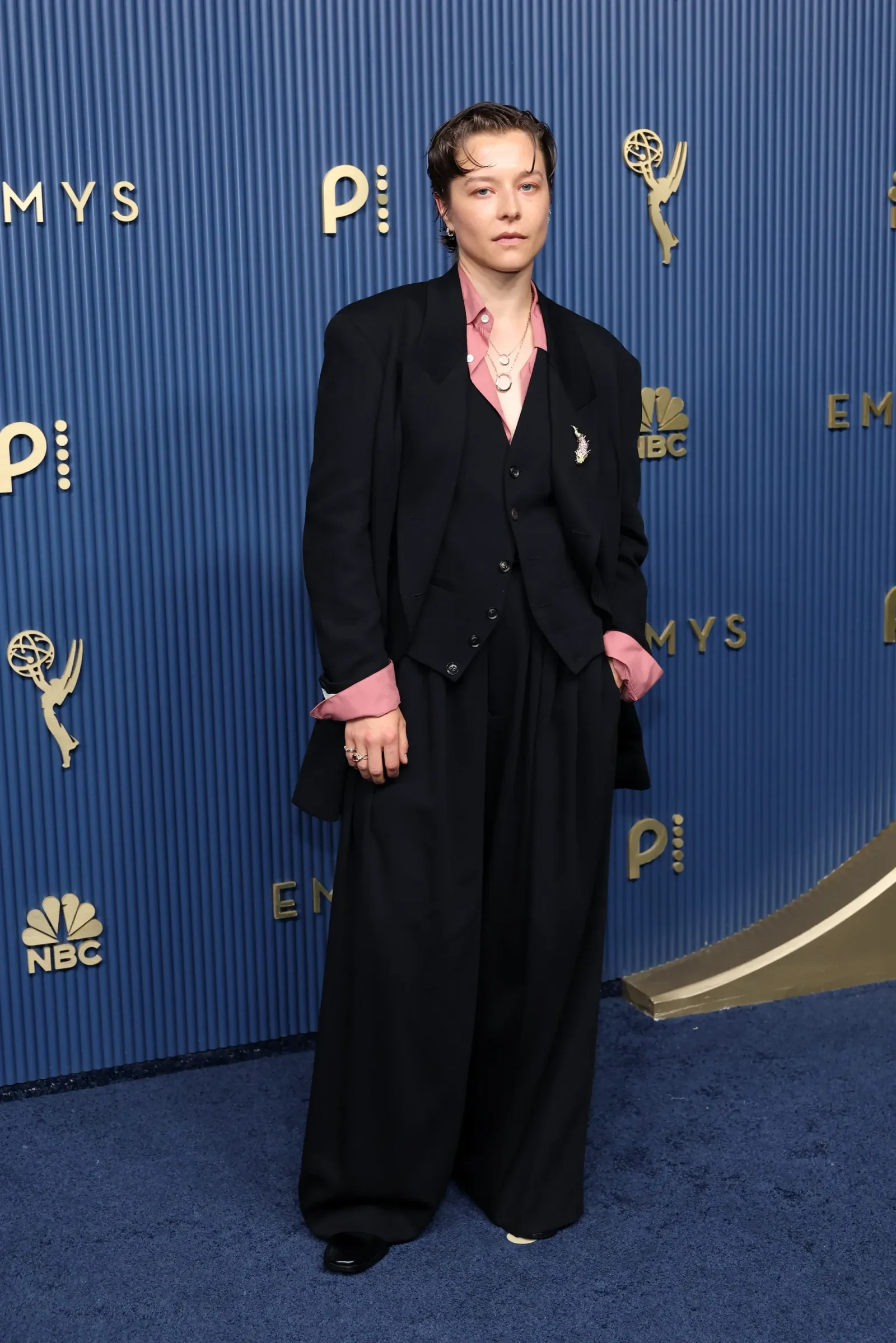 Person posing on a blue Emmy backdrop wearing a black suit with pink shirt cuffs and layered necklaces, accessories visible on lapel and fingers.