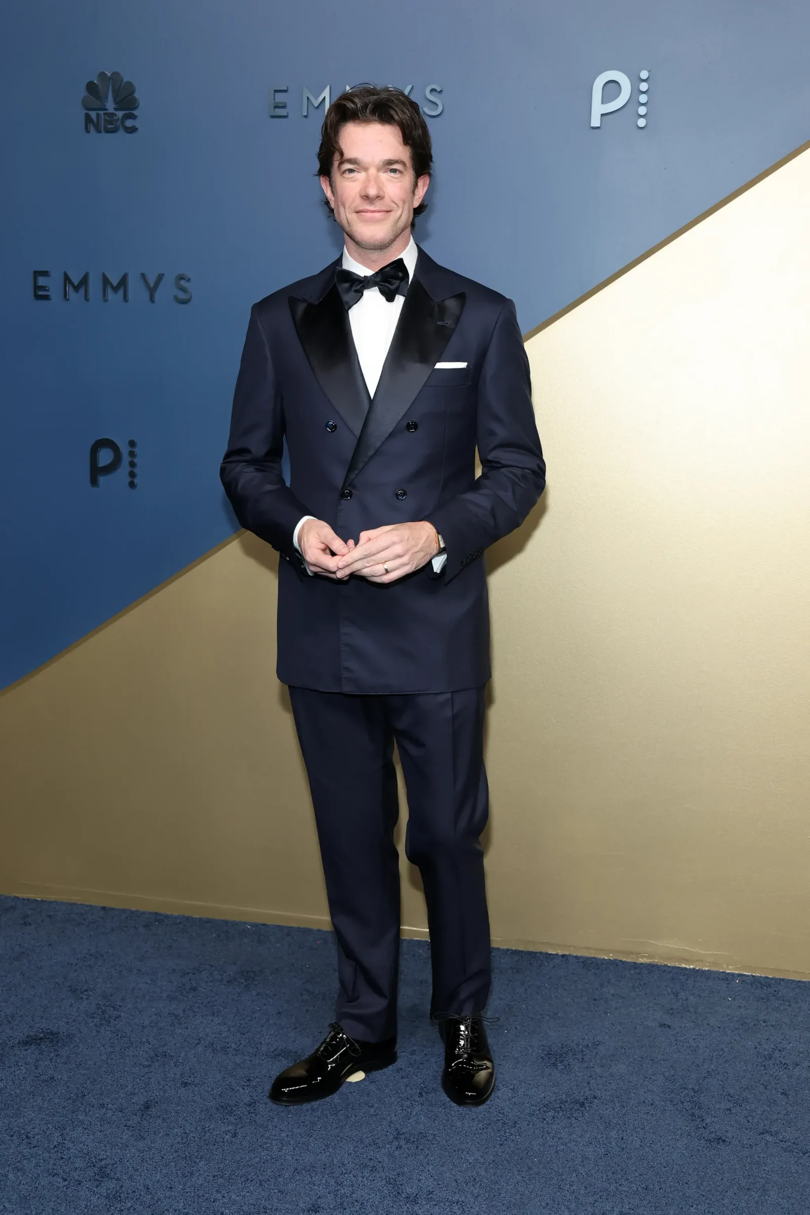 Man in a navy double-breasted tuxedo with a black bow tie and white pocket square, posing on a blue-and-gold Emmy Awards backdrop with hands clasped together.