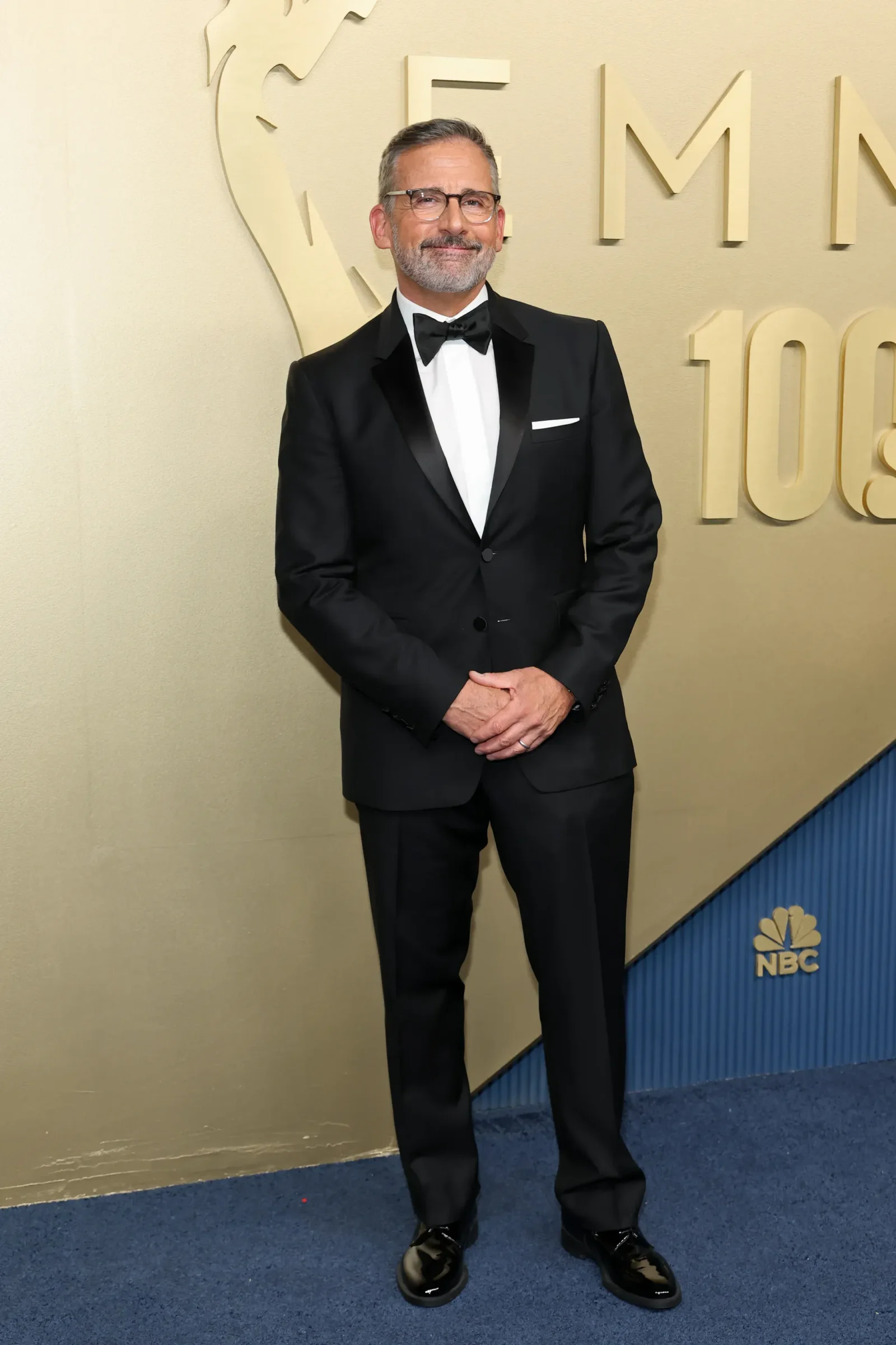 Man in a black tuxedo with bow tie stands smiling in front of a beige wall with large event branding at NBC.