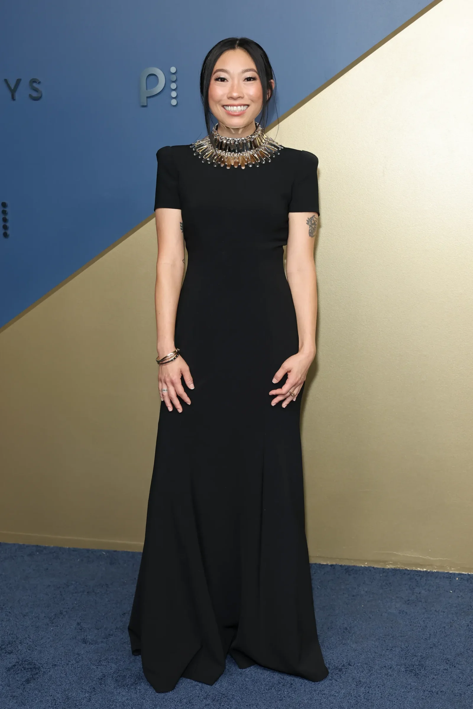 Woman in a long black dress wearing a large metallic necklace, smiling at the camera against a blue and gold diagonal wall backdrop.