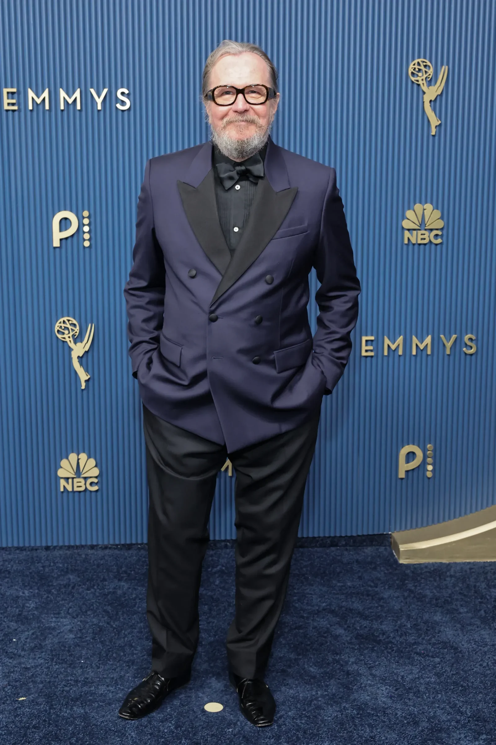 Man in a navy tuxedo with black lapels and bow tie posing on a blue Emmy Awards backdrop with NBC logos behind him