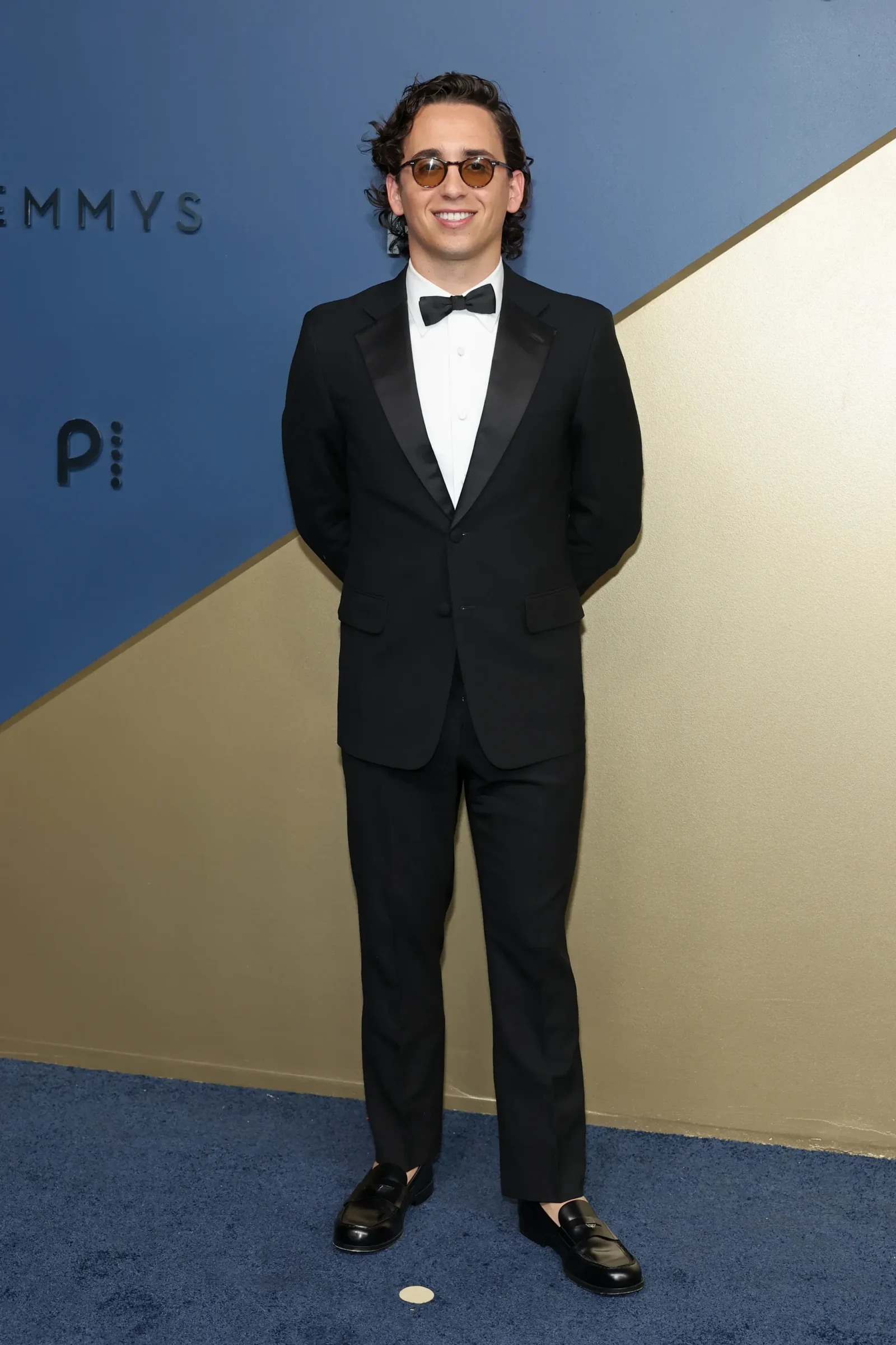 Man in a black tuxedo with white shirt and bow tie, wearing round sunglasses, posing for a photo against a blue and beige diagonal backdrop.