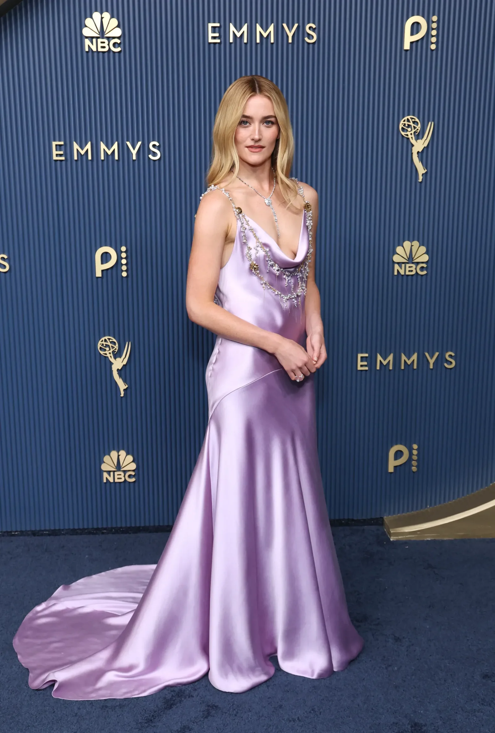 Blonde woman in a lavender satin gown with jeweled detailing posing on a blue Emmy backdrop.