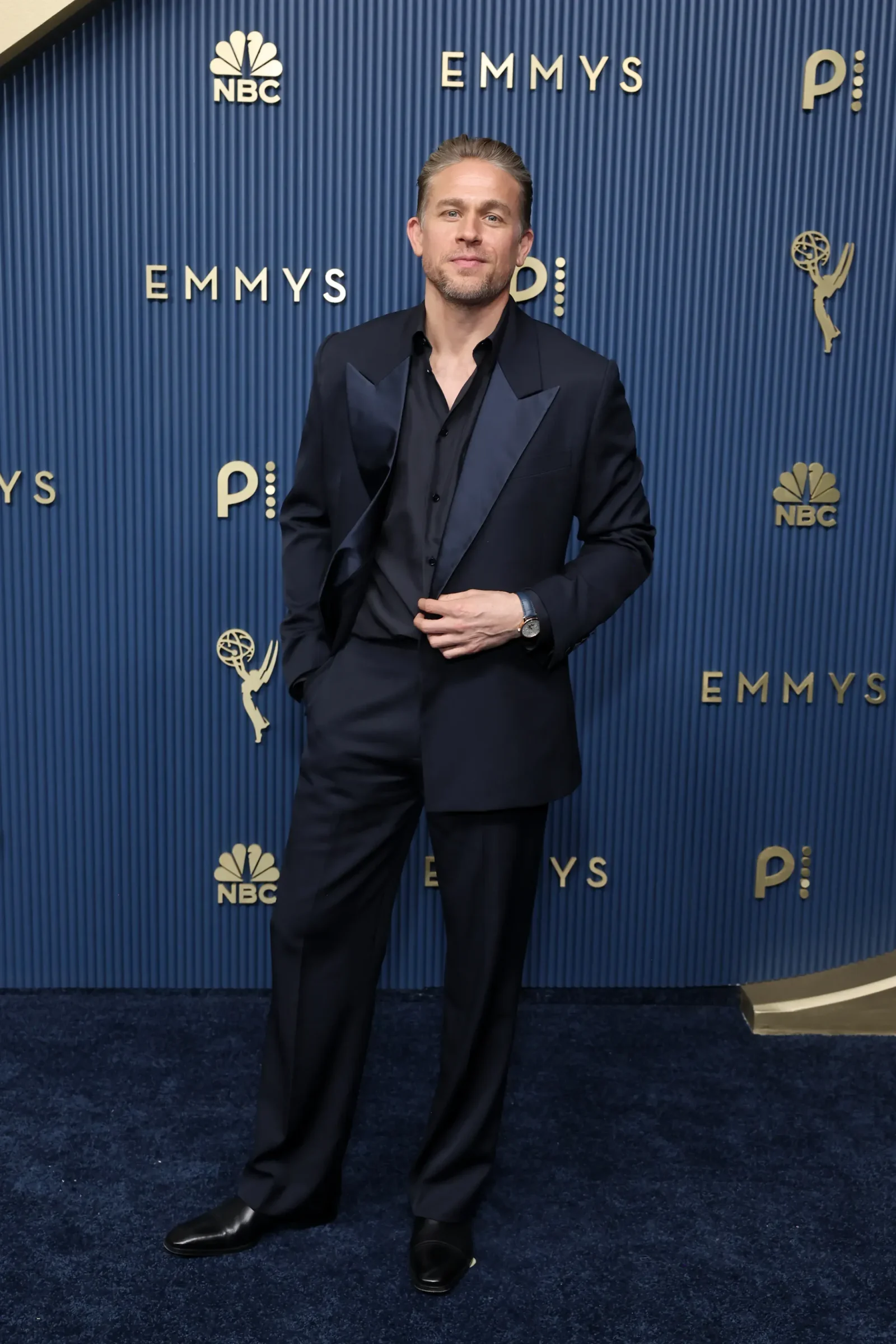 Man in a dark suit posing for photos on a blue Emmy Awards backdrop with NBC logos nearby in the background, full body visible.
