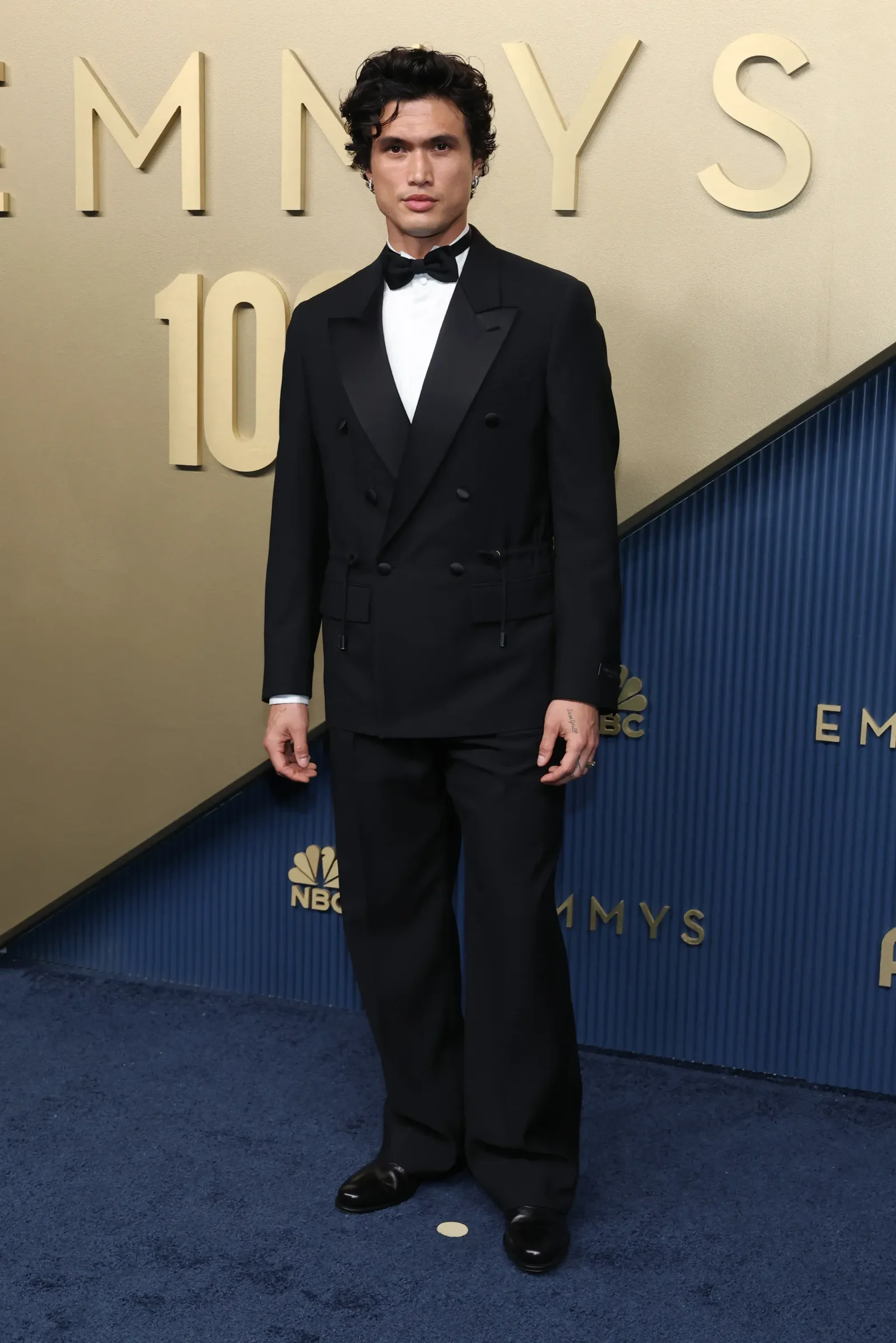 Man in a black double-breasted tuxedo with a bow tie standing on a blue carpet at a red carpet event, with large gold EMmys letters in the background behind him.
