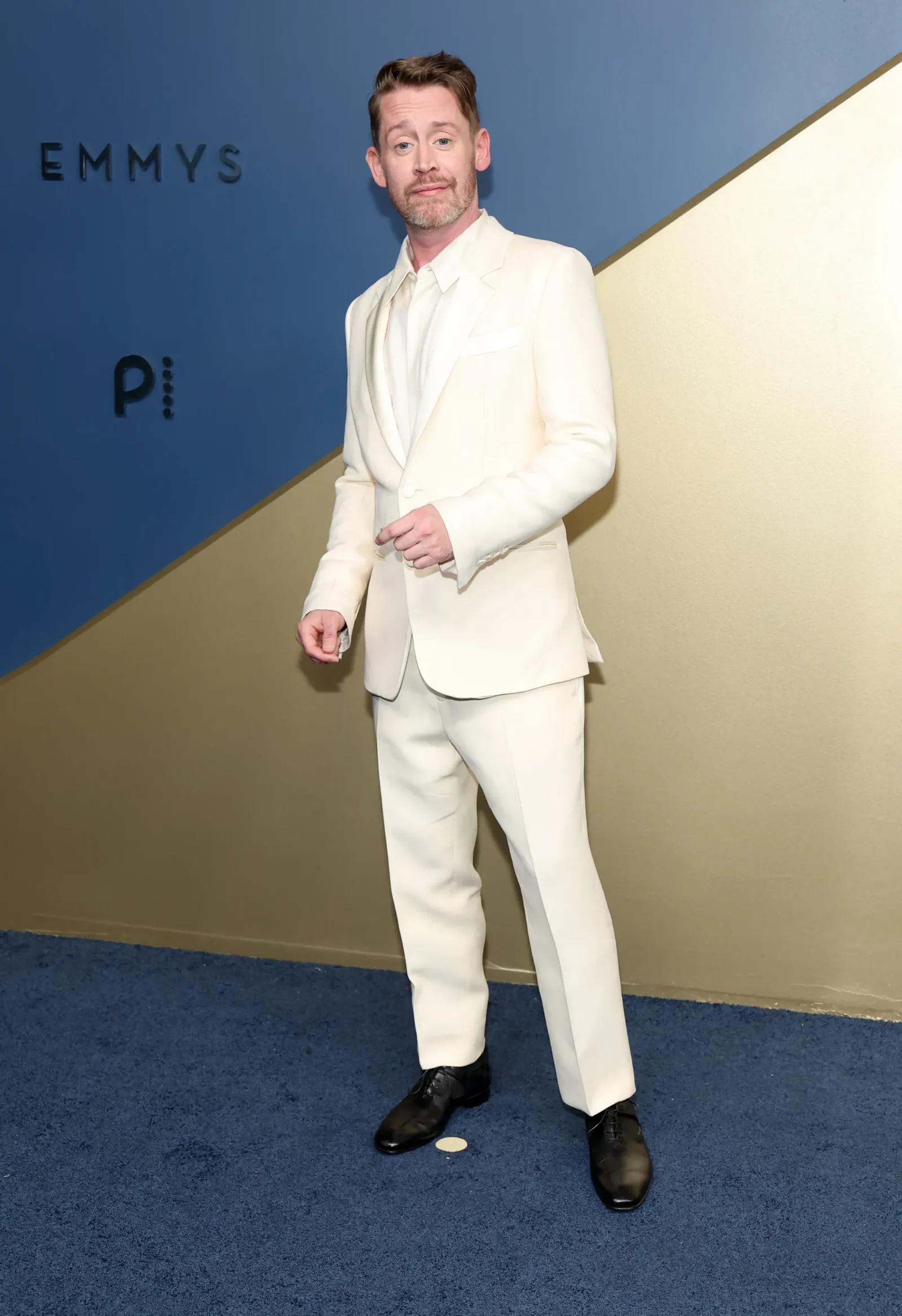 Man in a white/cream suit posing for a photo at an event, standing on a blue carpeted floor with a blue and beige backdrop.