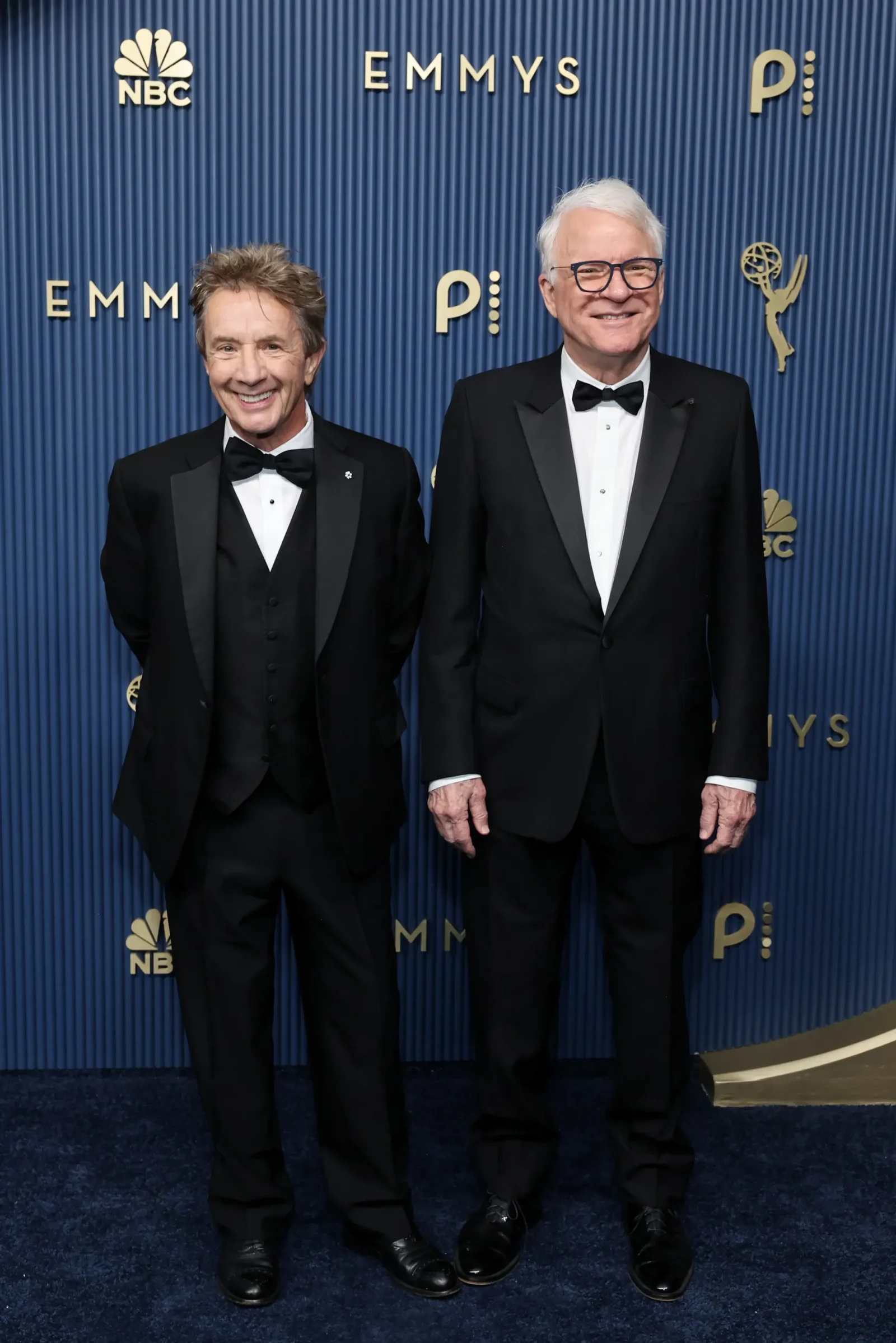 Two smiling men in black tuxedos stand side by side on a blue carpet at an Emmy Awards backdrop featuring NBC branding and gold lettering/graphics.