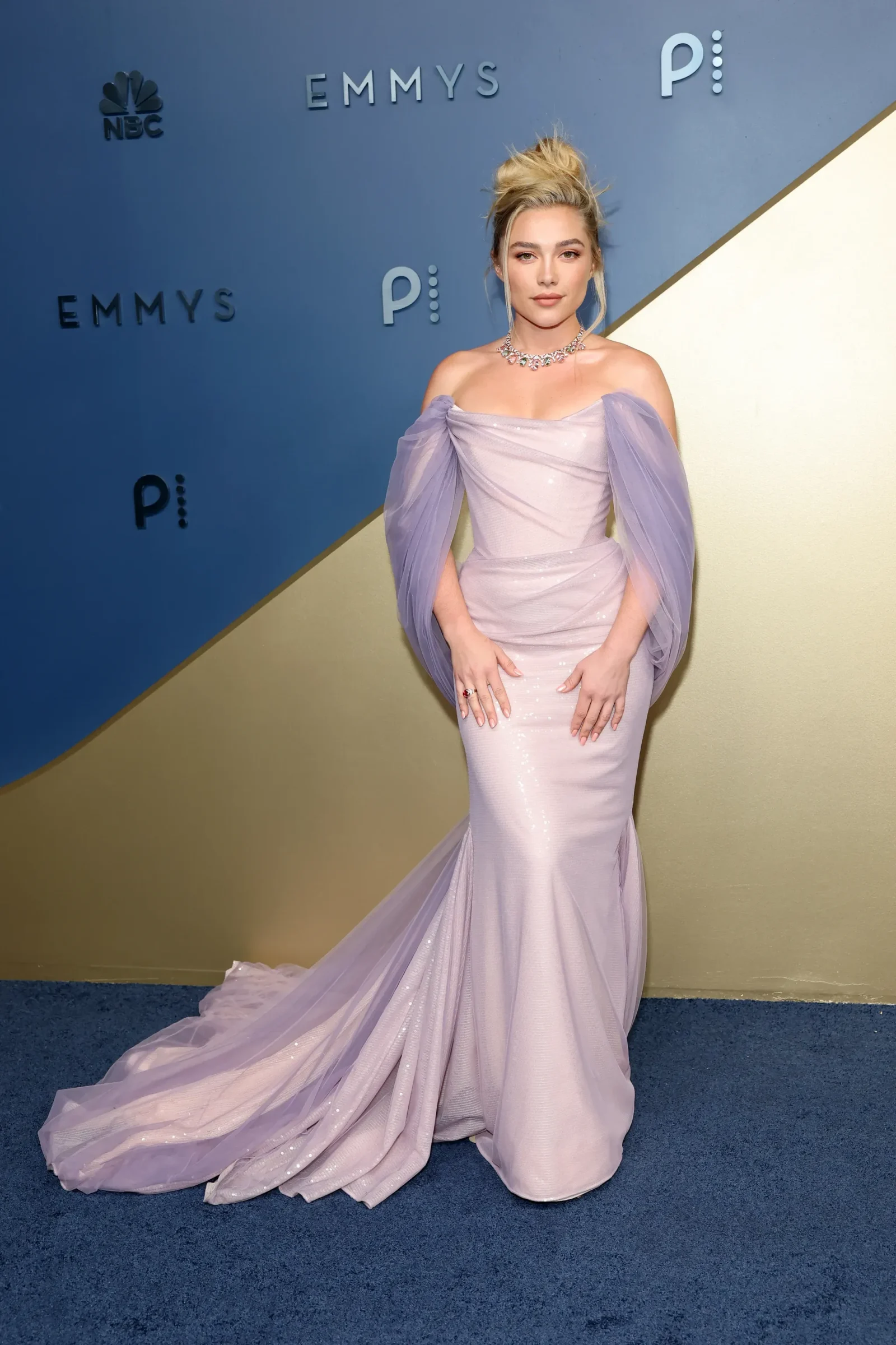 Woman in a lavender off-shoulder evening gown with draped sleeves posing on a blue-and-gold step-and-repeat backdrop at an Emmy Awards event.