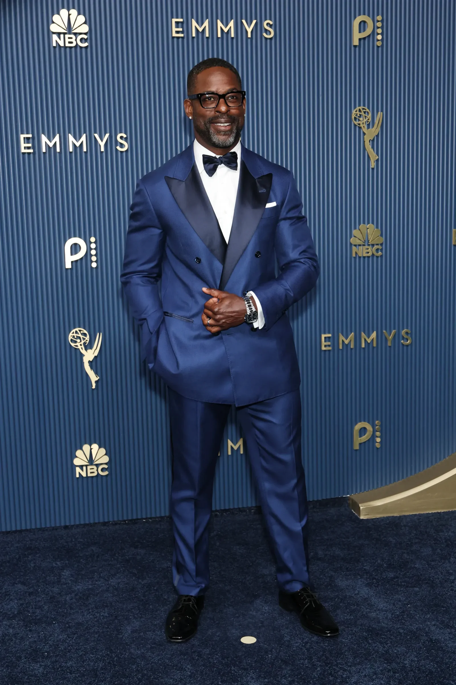 Man in a blue tuxedo with a black bow tie posing on a blue Emmy Awards backdrop with NBC logos nearby.