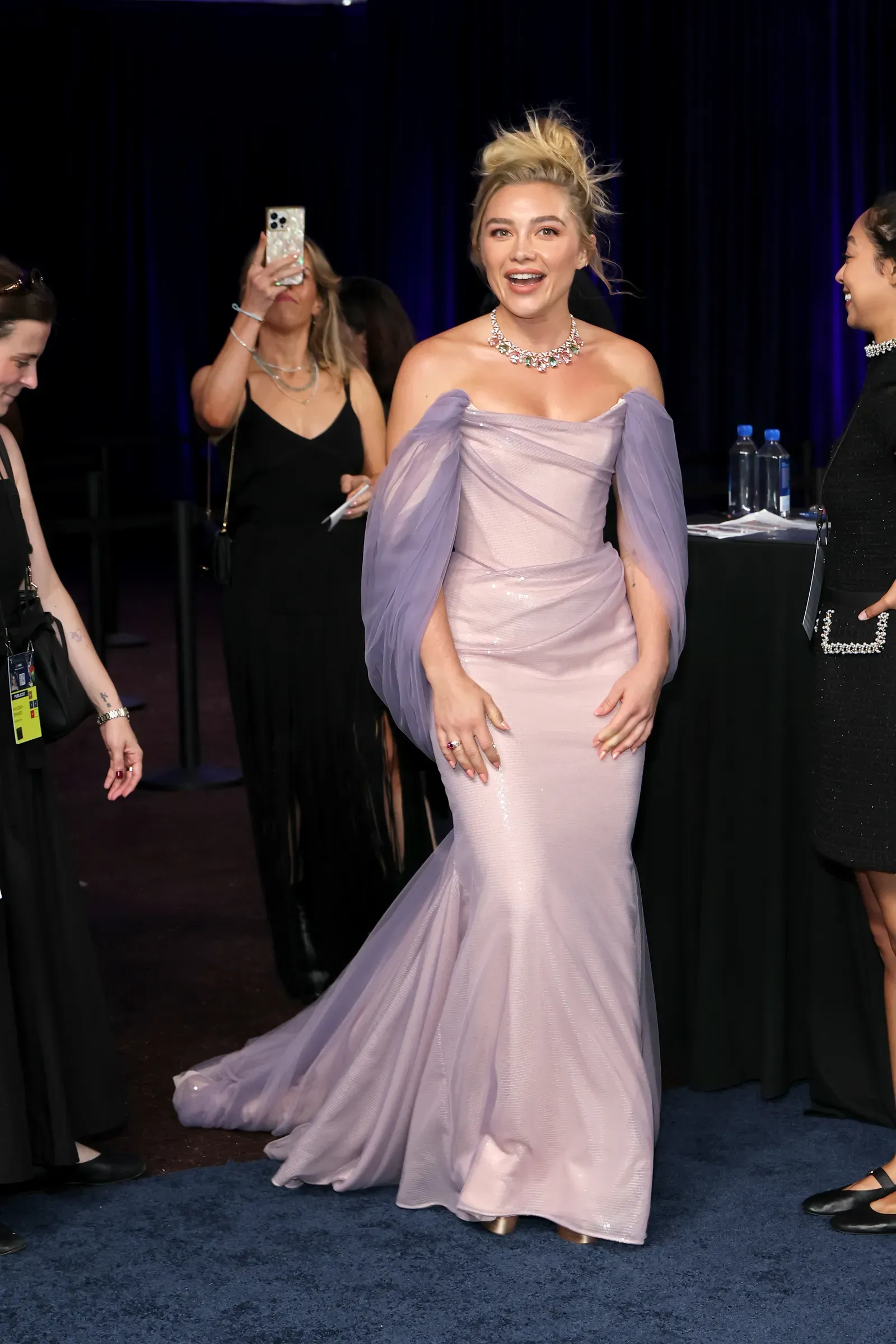Woman in a lavender off-the-shoulder gown posing on a blue carpet at a formal event, others nearby taking photos and talking in the background.
