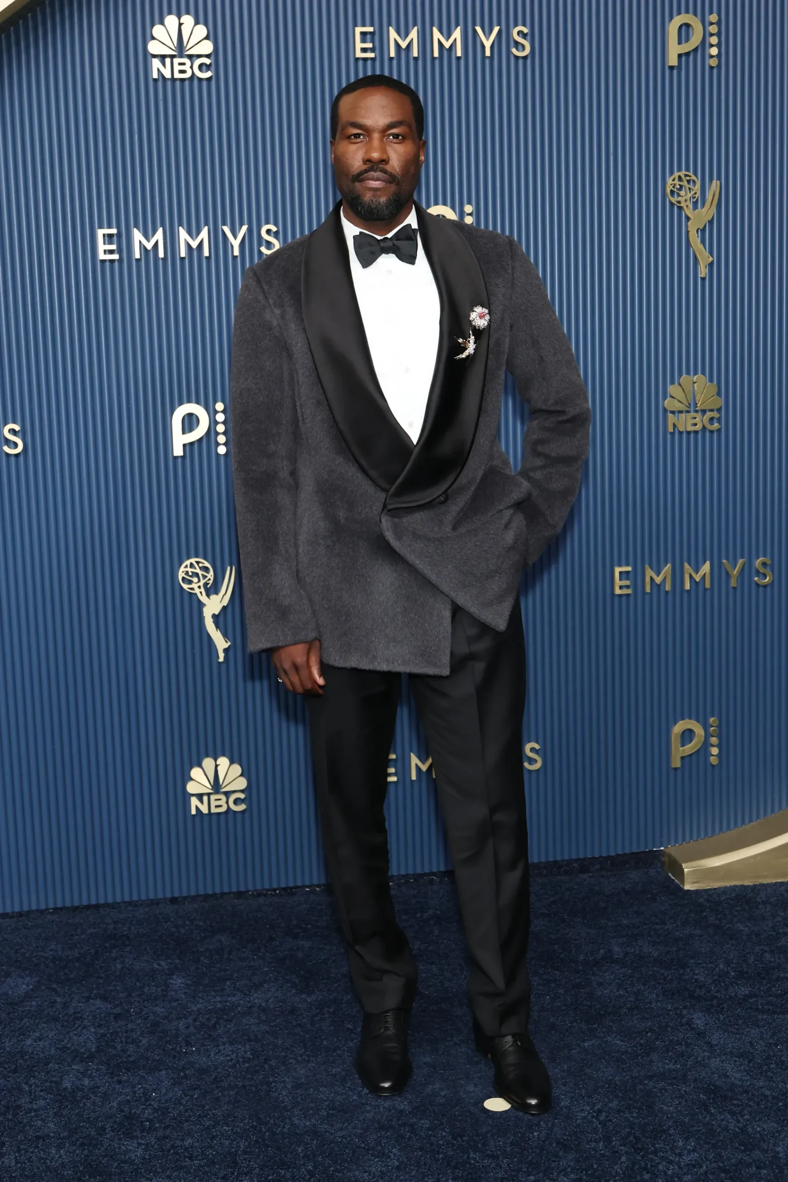African American man in a black tuxedo with white shirt and bow tie posing on a blue Emmy Awards backdrop with NBC logos.