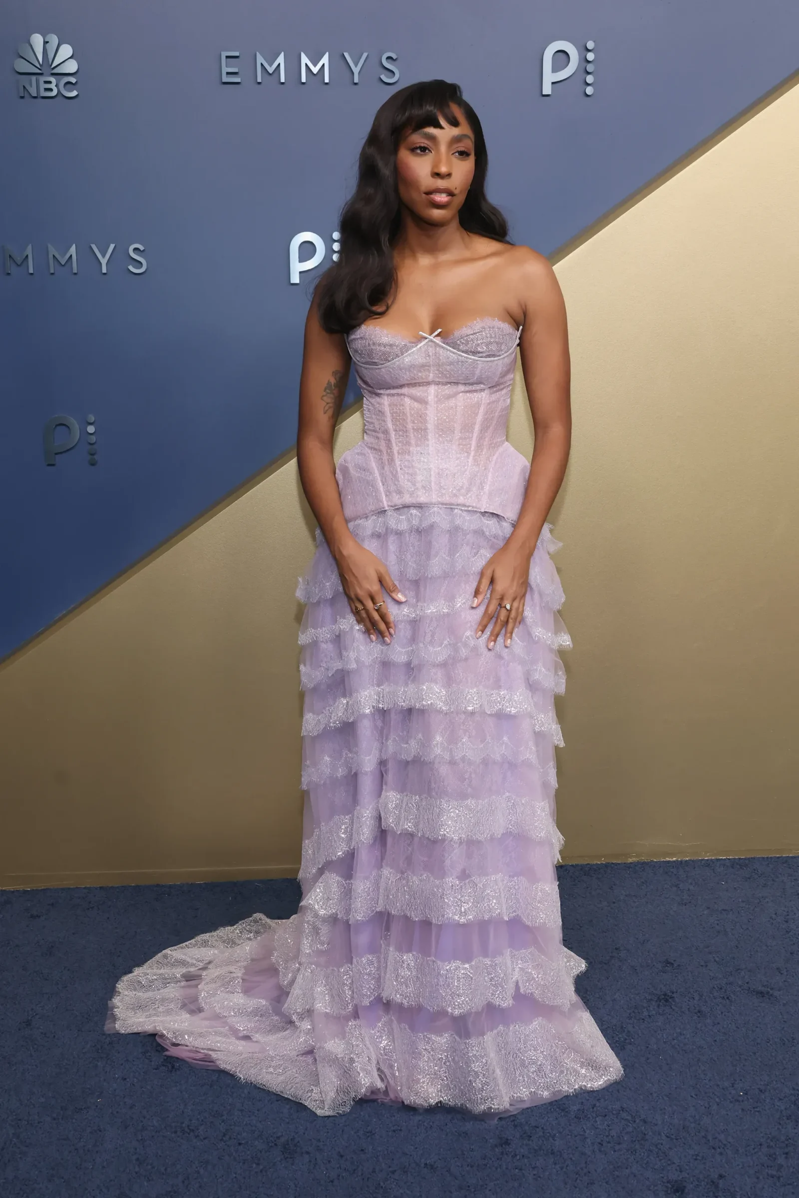 Woman in a lavender strapless lace gown with a layered train on a blue carpet at an Emmy event on a backdrop wall.