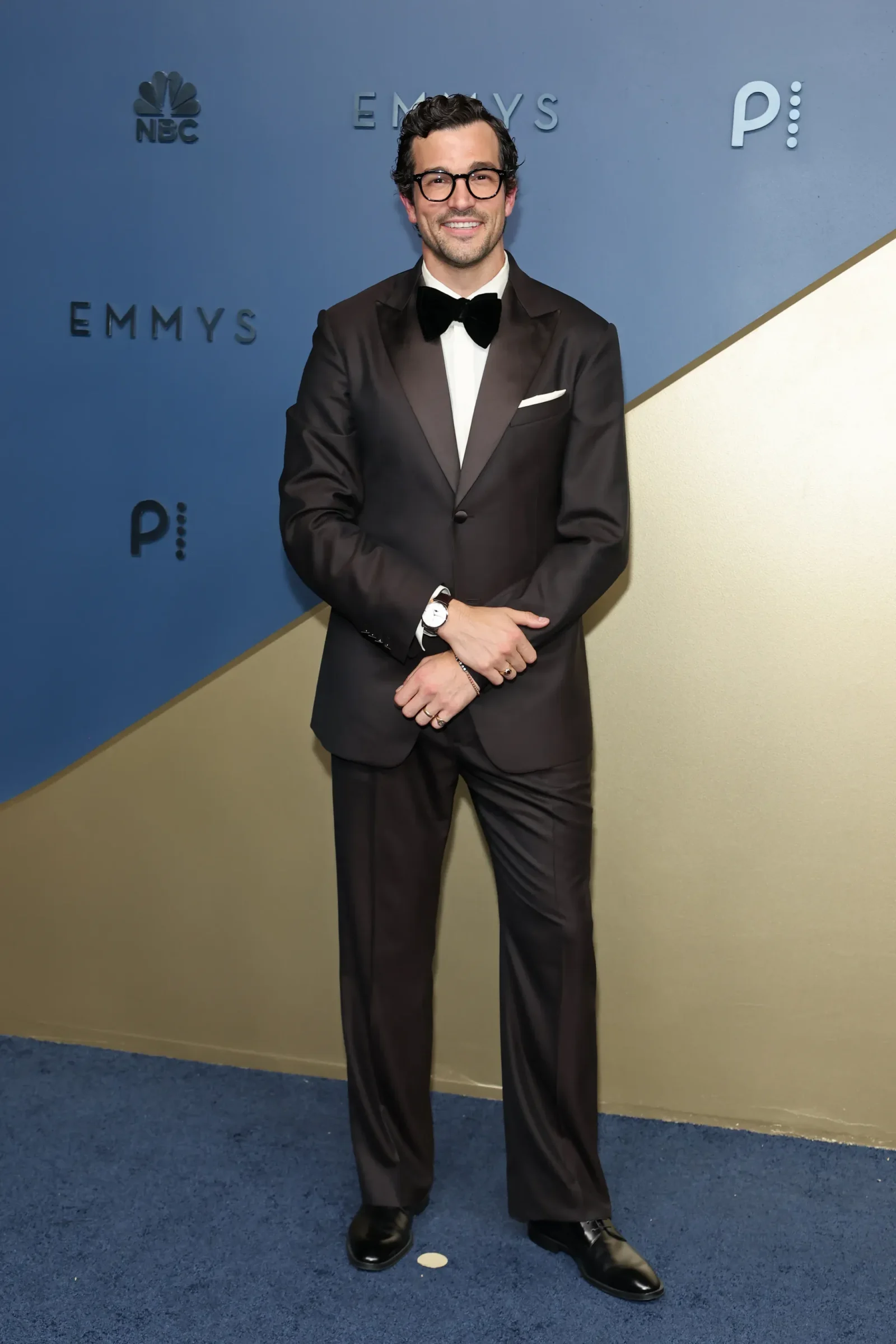 Man in a black tuxedo with a bow tie and glasses posing against a blue Emmy Awards backdrop.