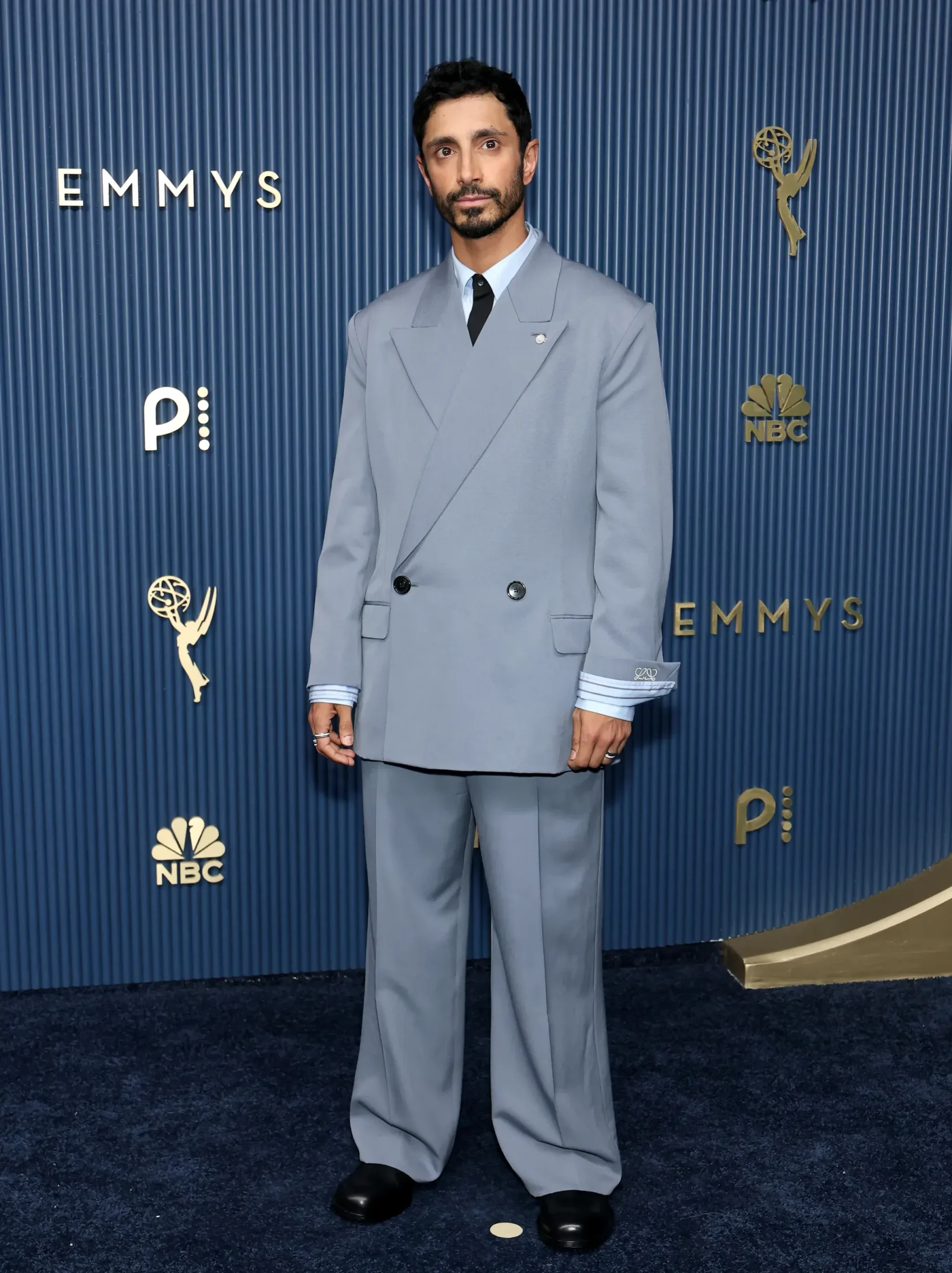 Man posing on a blue Emmy Awards backdrop in a light gray double-breasted suit, black tie, and black shoes.