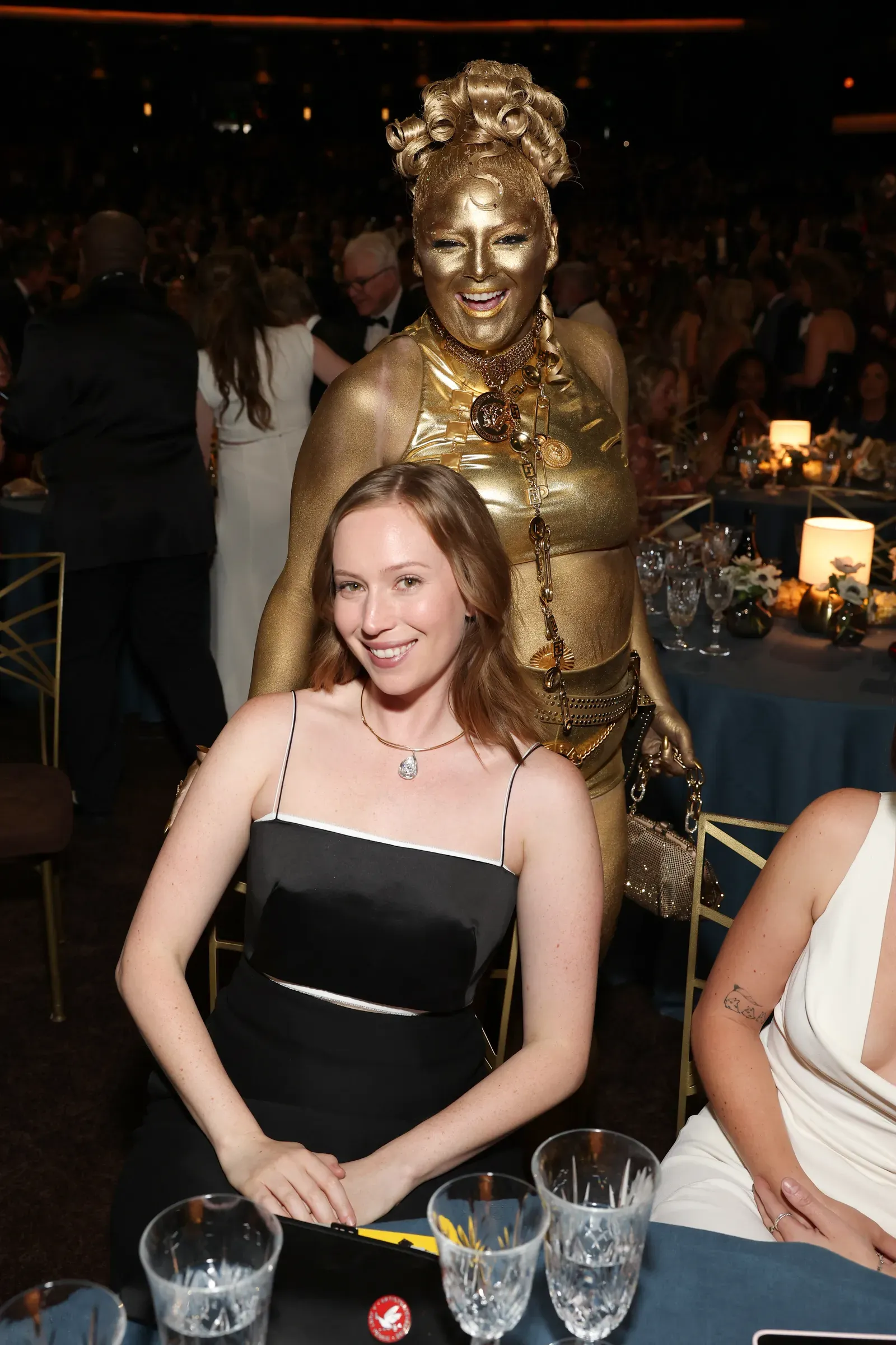 Woman with gold body paint and sculpted hair standing behind a seated woman in a black dress at a formal event, both smiling at the camera.
