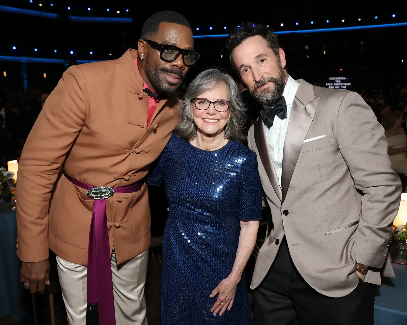 Three people pose for a photo at a formal event: a man in a tan jacket with a magenta sash, a gray-haired woman in a blue sequined dress, and a man in a light tan tuxedo with a black bow tie, all smiling at the camera.
